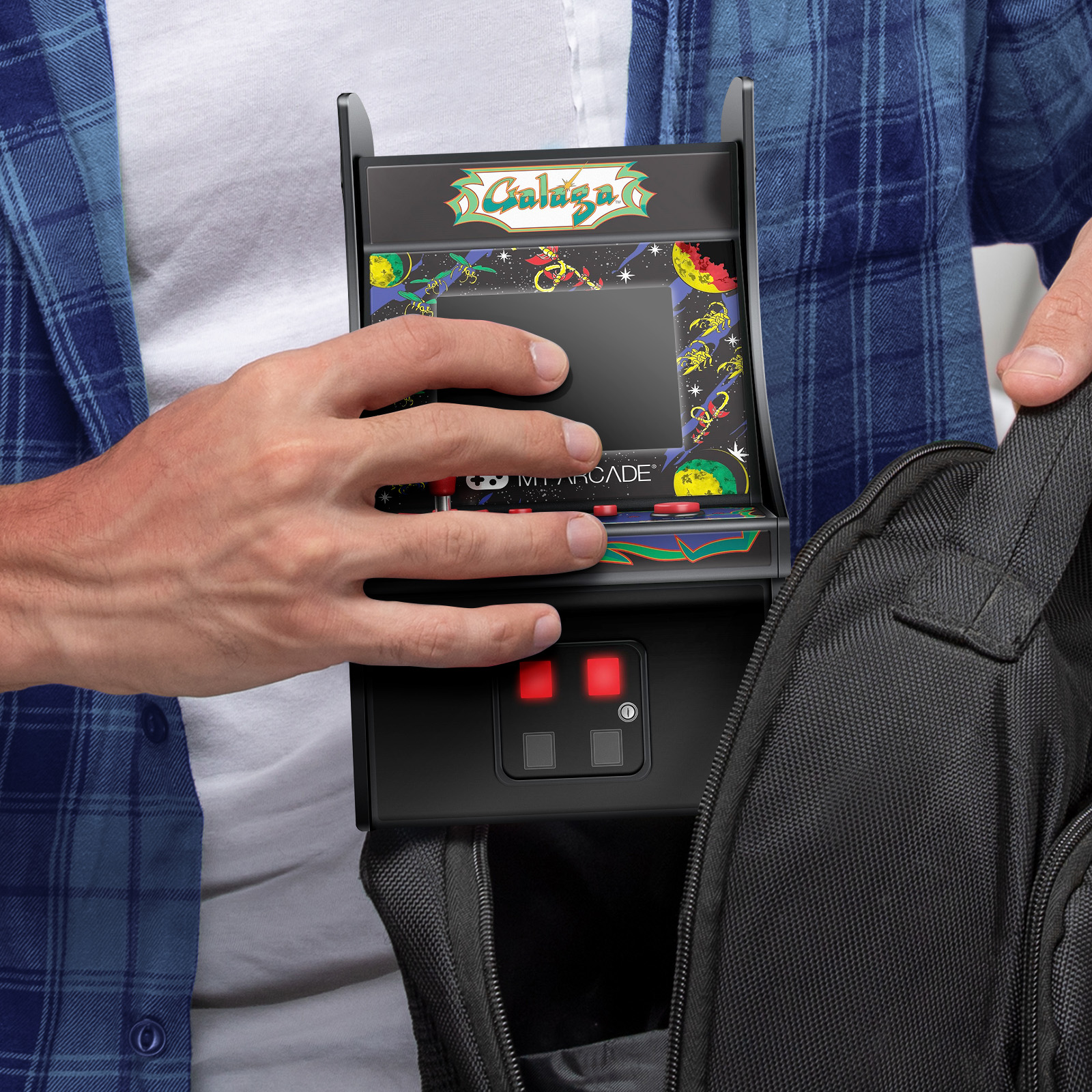 Una persona tiene in mano un mini gioco arcade, mettendolo in una borsa nera. Il gioco presenta il logo Galaga e pulsanti rossi.