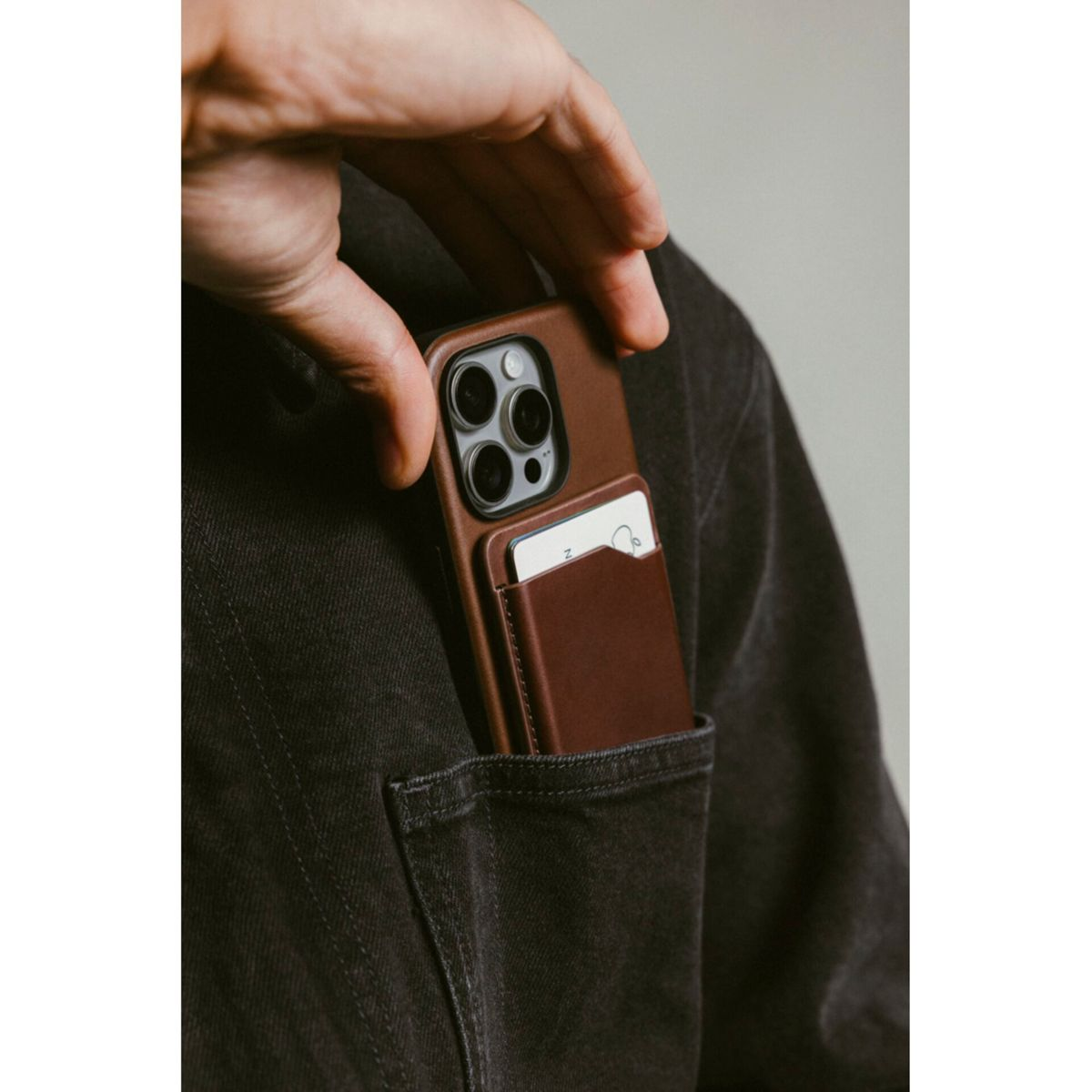Una mano coloca una funda marrón para teléfono con un tarjetero en un bolsillo de una chaqueta vaquera negra. La funda tiene un teléfono.