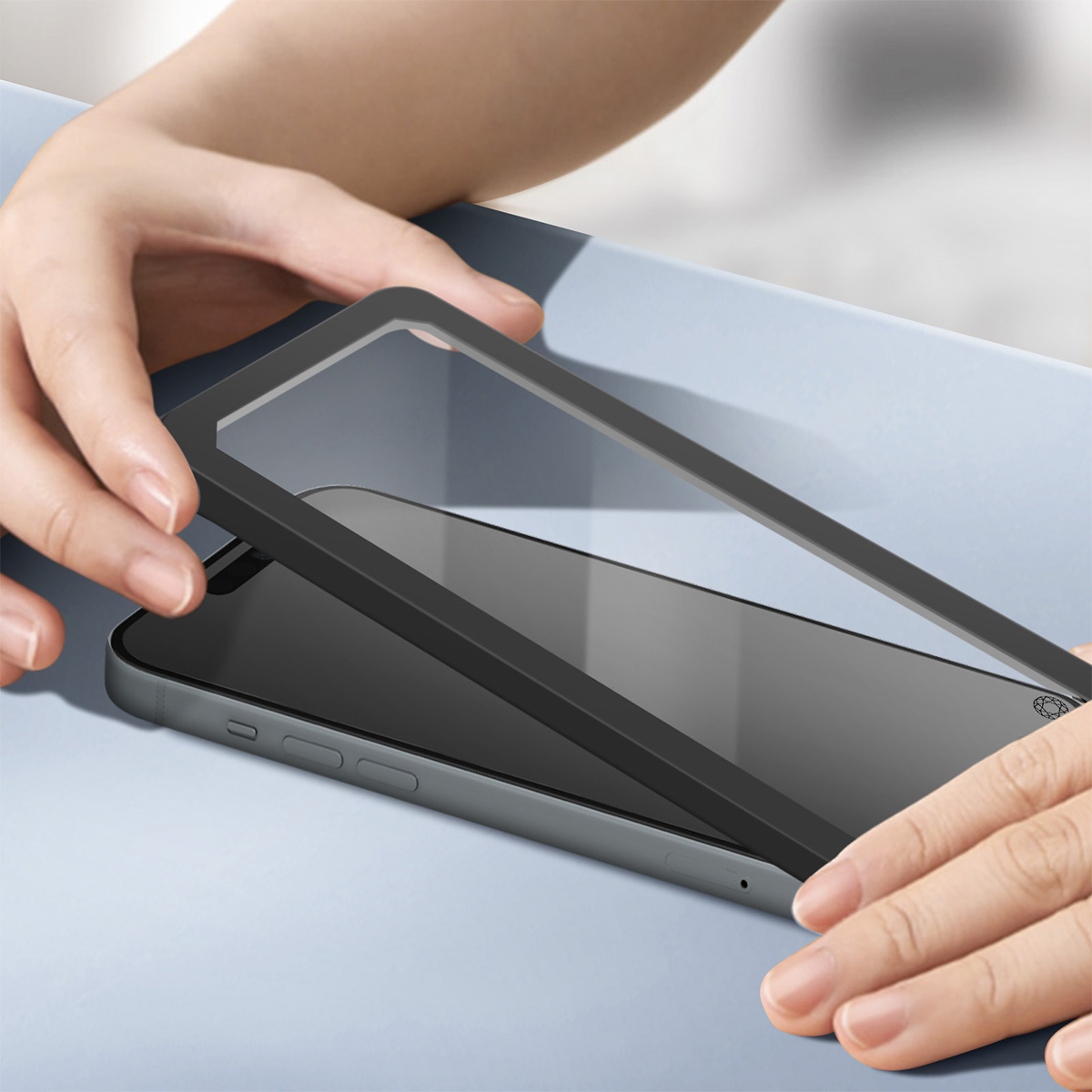 Le mani posizionano una cornice nera su uno smartphone grigio. La cornice ha una protezione schermo trasparente. Lo sfondo è azzurro.