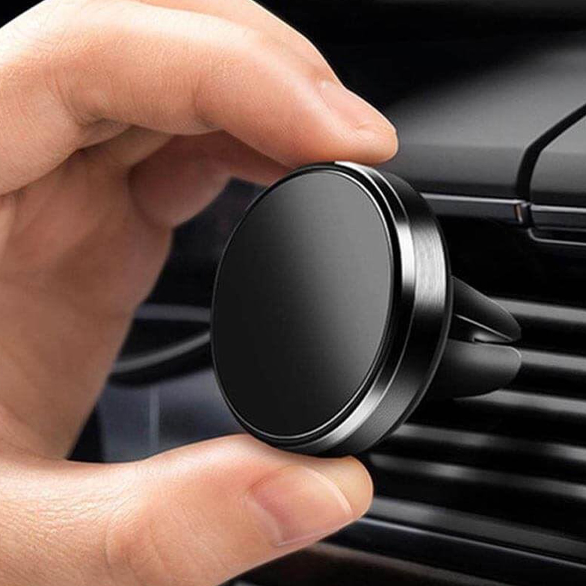 Una mano tiene un supporto nero per bocchetta dell'auto. Il supporto è rotondo con finiture argentate. Lo sfondo mostra le bocchette dell'auto.