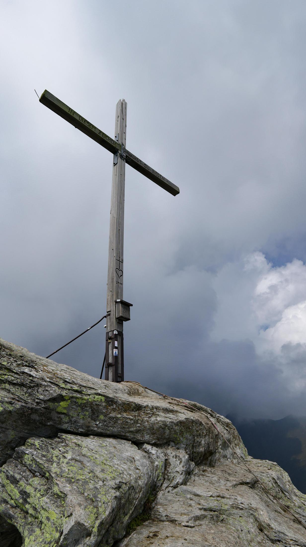 Croce di legno su una cima rocciosa sotto un cielo nuvoloso. La croce ha delle iscrizioni. In primo piano rocce muschiose.