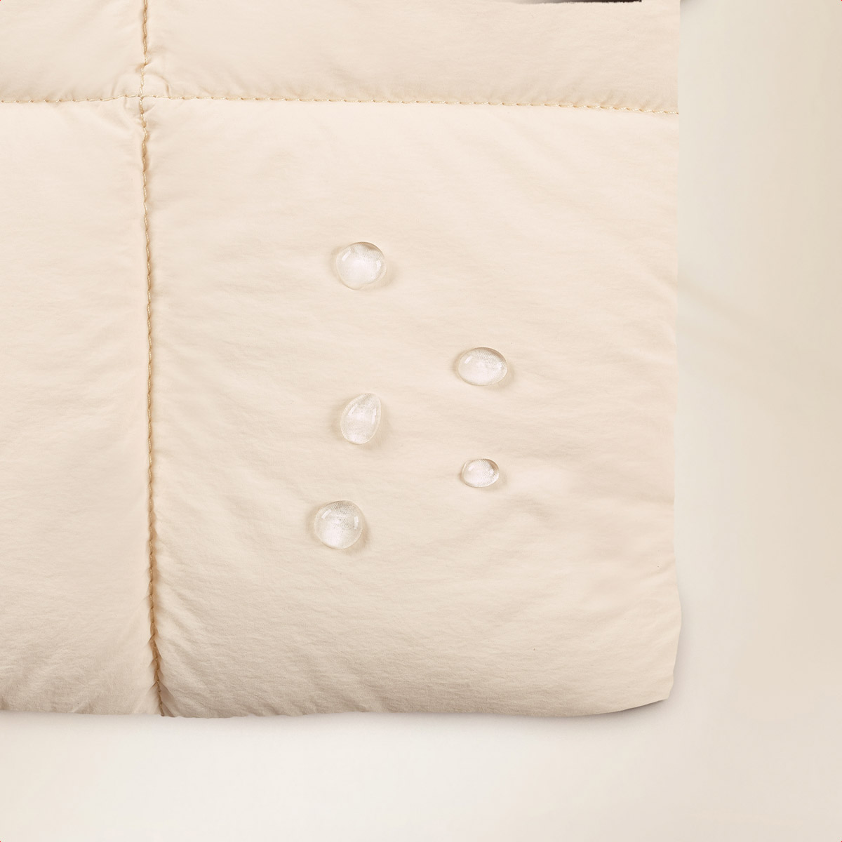 Close-up of beige fabric with water droplets. Fabric is quilted. Drops are clear and spherical. Background is pale beige.