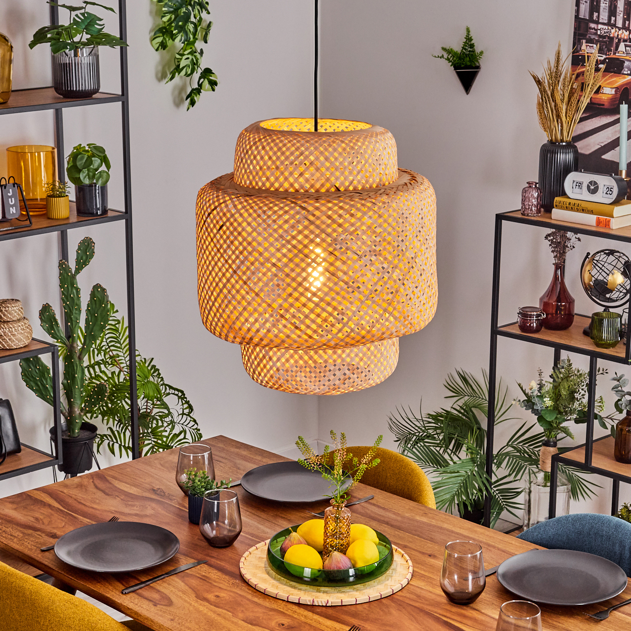 Ein Esstisch mit Tellern, Gläsern und einer Obstschale unter einer gewebten Lampe. Regale zeigen Pflanzen und Dekoration.