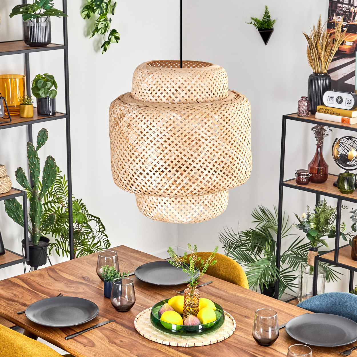 Ein Esstisch mit Tellern, Gläsern und einem Obst-Mittelstück. Eine gewebte Lampe hängt darüber. Regale mit Pflanzen im Hintergrund.