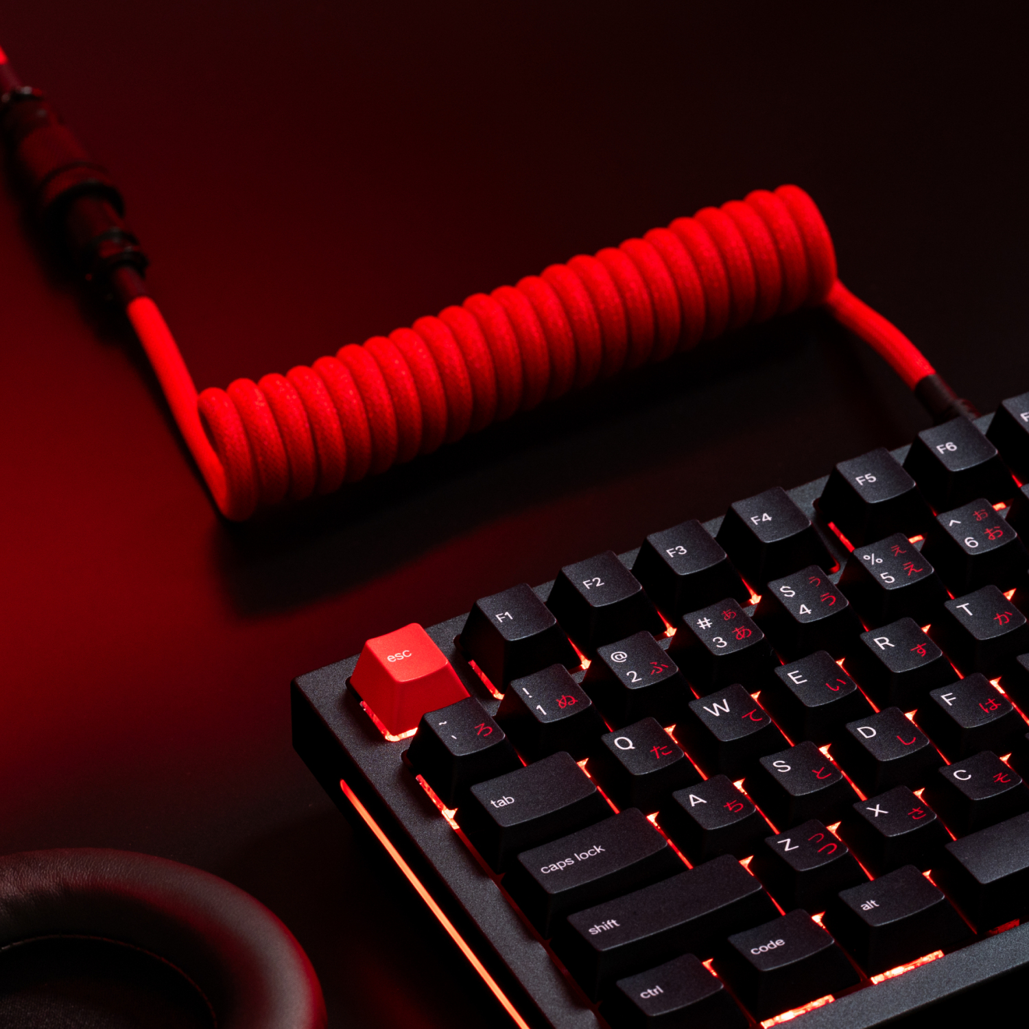Primo piano di una tastiera meccanica con cavo a spirale rosso. La tastiera è nera con tasti retroilluminati rossi. Tasto ESC rosso prominente.