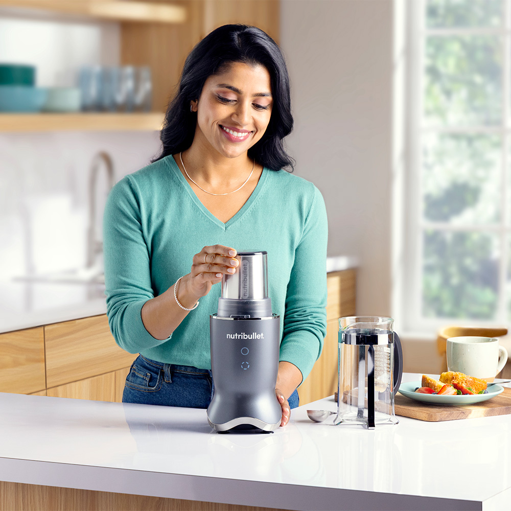 A woman smiles while assembling a Nutribullet blender on a white countertop. Breakfast is set out in the background.