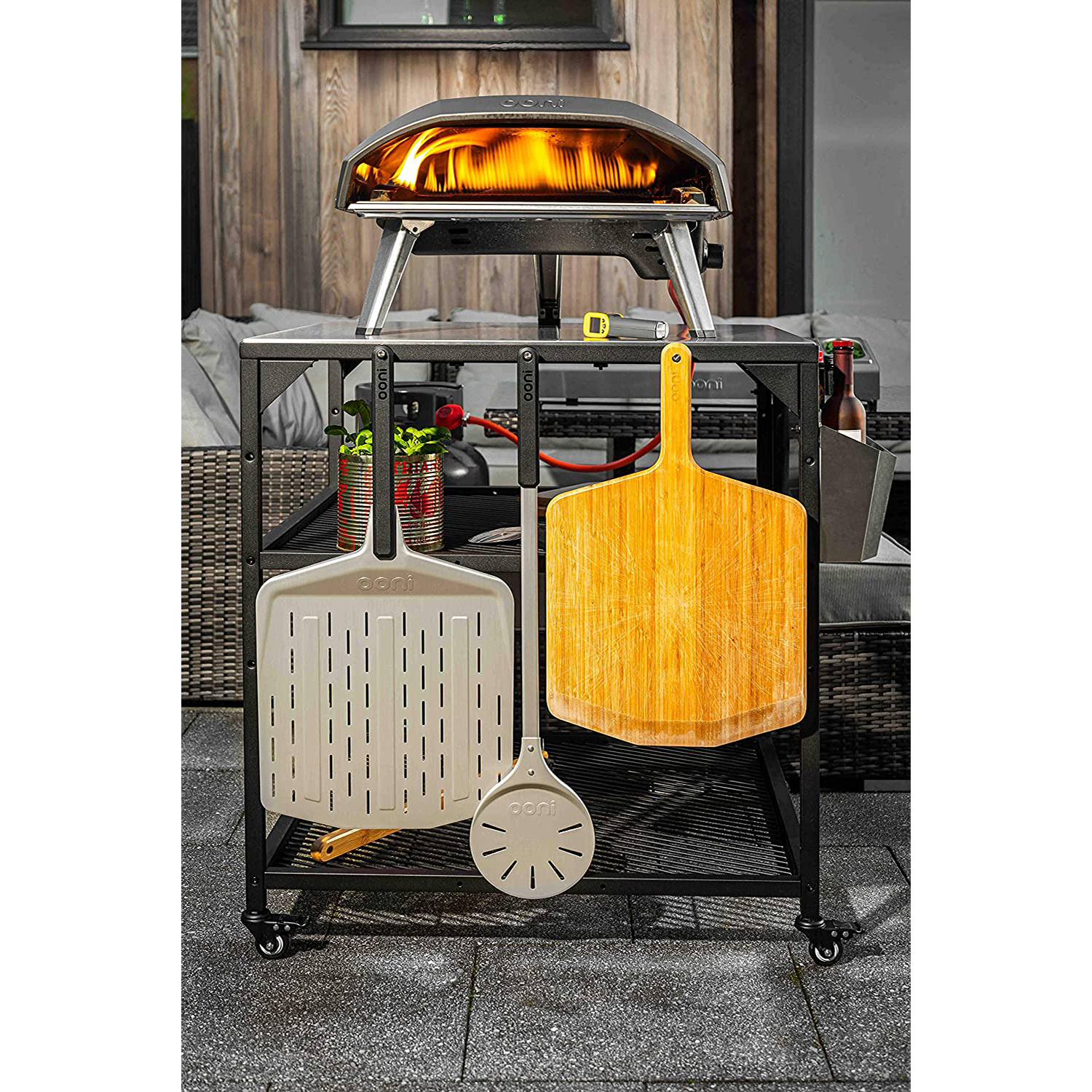 Un forno per pizza con fiamme, con pala per pizza in legno e attrezzi. Su un carrello di metallo nero, con parete in legno sullo sfondo.