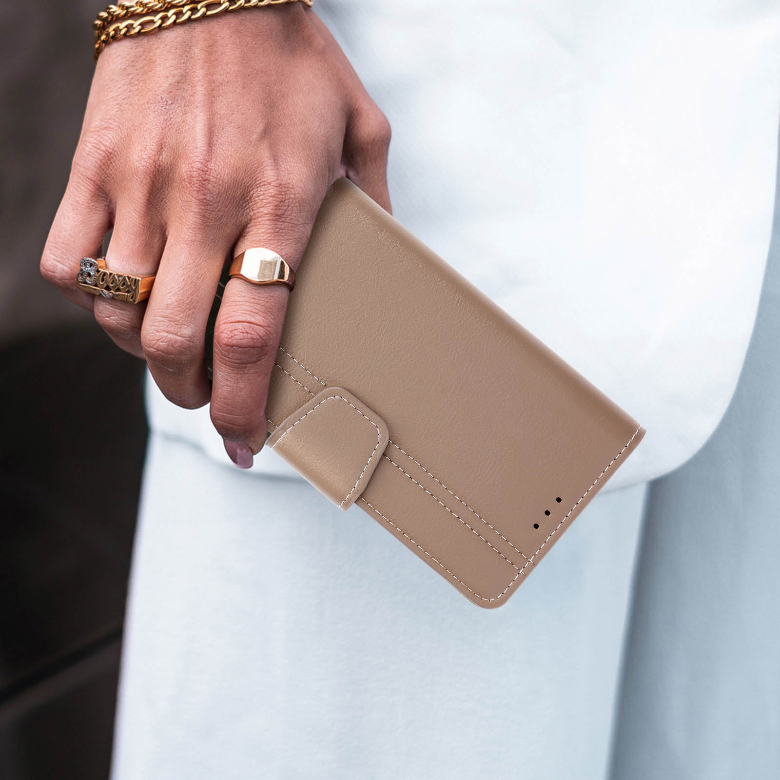 Mano che tiene una custodia beige con anelli d'oro e un braccialetto. Abito bianco sullo sfondo.