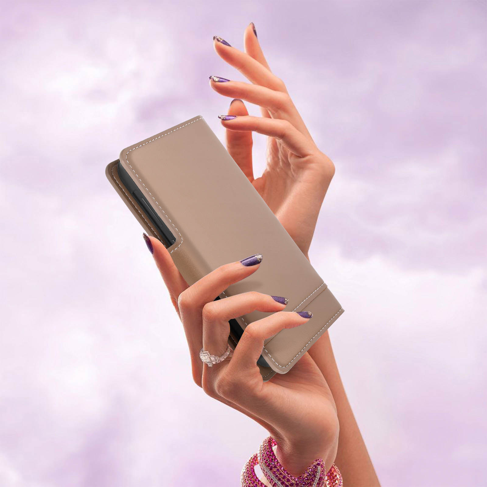 Mani che tengono una custodia beige per telefono su sfondo viola. Unghie dipinte di viola. Un anello e un braccialetto sono visibili.