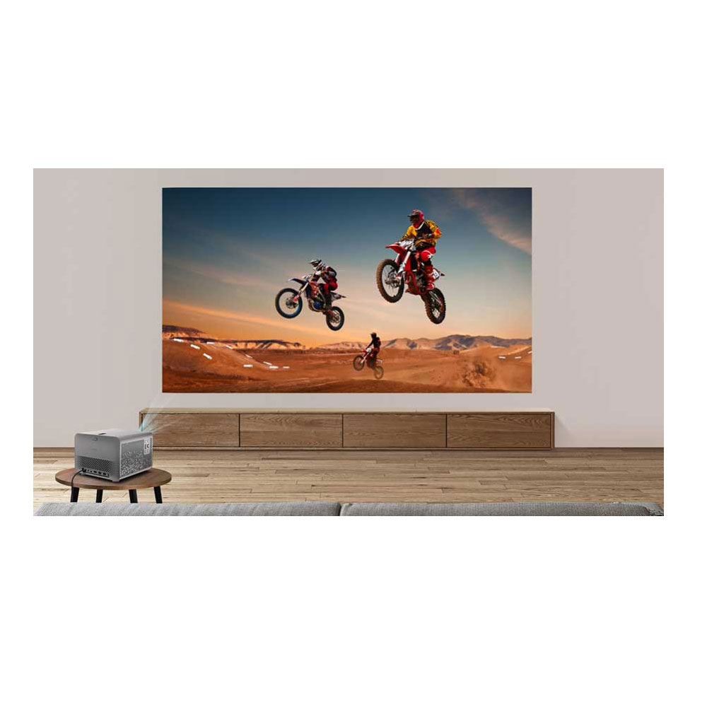 Un projecteur affiche une scène de motocross avec trois pilotes sautant en l'air. La pièce a des meubles en bois et un sol brun.