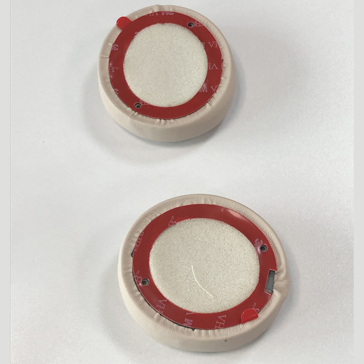 Twee beige oorkussens voor hoofdtelefoons met rode kleefringen en witte schuimcentra op een wit oppervlak.