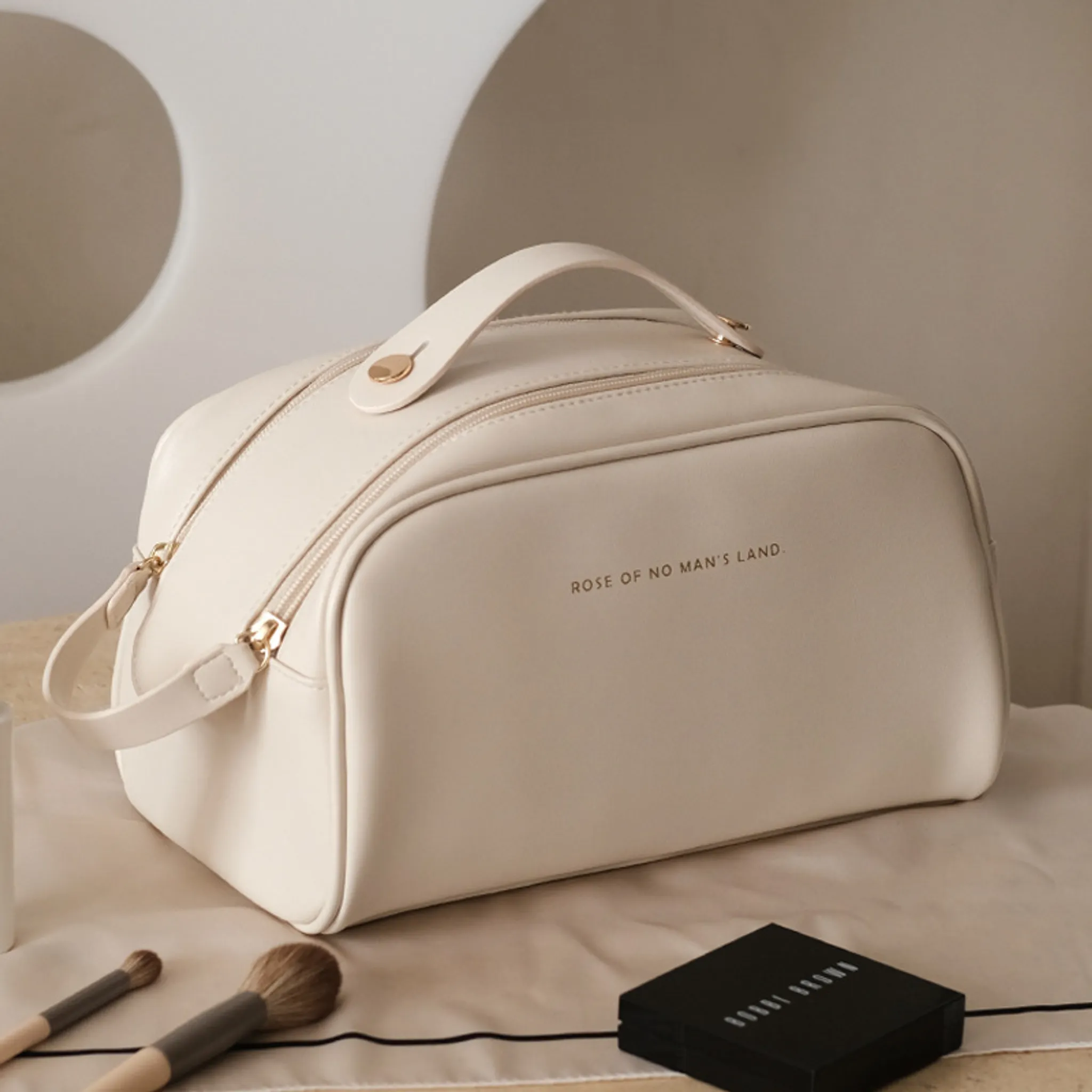 Eine cremefarbene Kosmetiktasche mit goldenen Akzenten liegt auf einem Tuch. Zwei Make-up-Pinsel und ein schwarzer Kompakt liegen daneben.