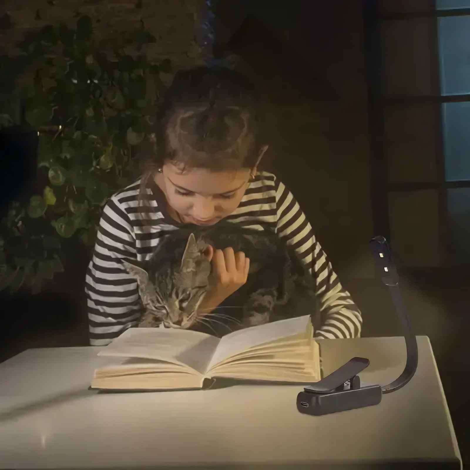 Ein Mädchen im gestreiften Hemd hält eine Katze, während es unter einer Klemmlampe ein aufgeschlagenes Buch liest.