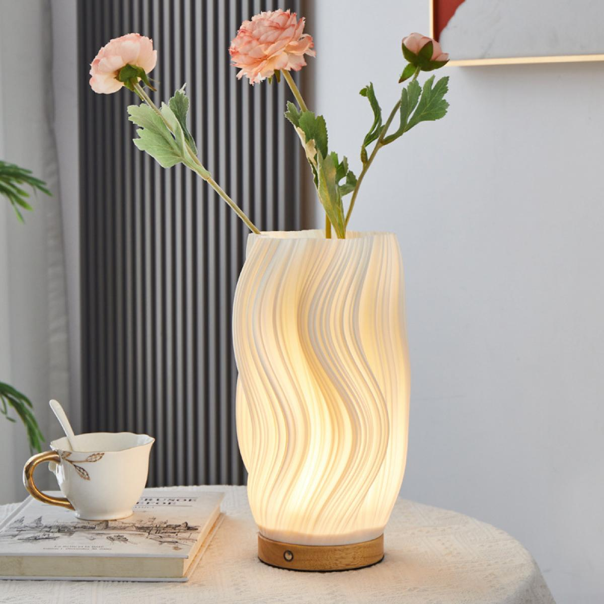 Eine wellenförmige weiße Vase mit rosa Blumen steht neben einem Buch und einer Tasse auf einem Tisch.