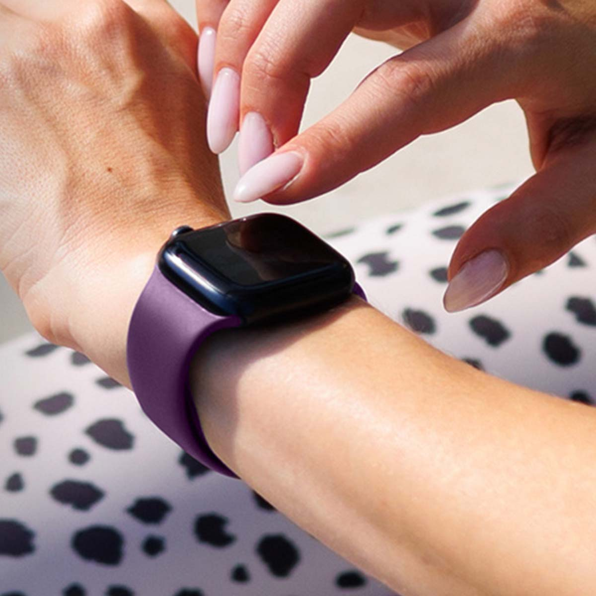 Primo piano di un polso con uno smartwatch viola. Dita con unghie rosa chiaro toccano il quadrante dell'orologio.