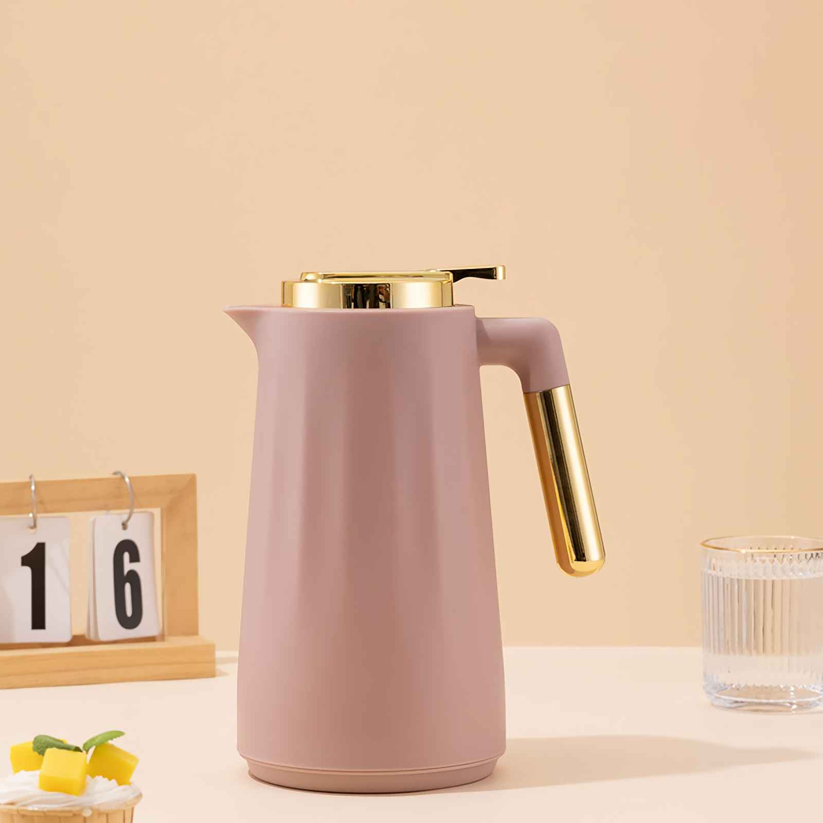 Ein staubrosafarbener Thermoskanne mit goldenen Akzenten steht neben einem Glas Wasser und einem Kalender.
