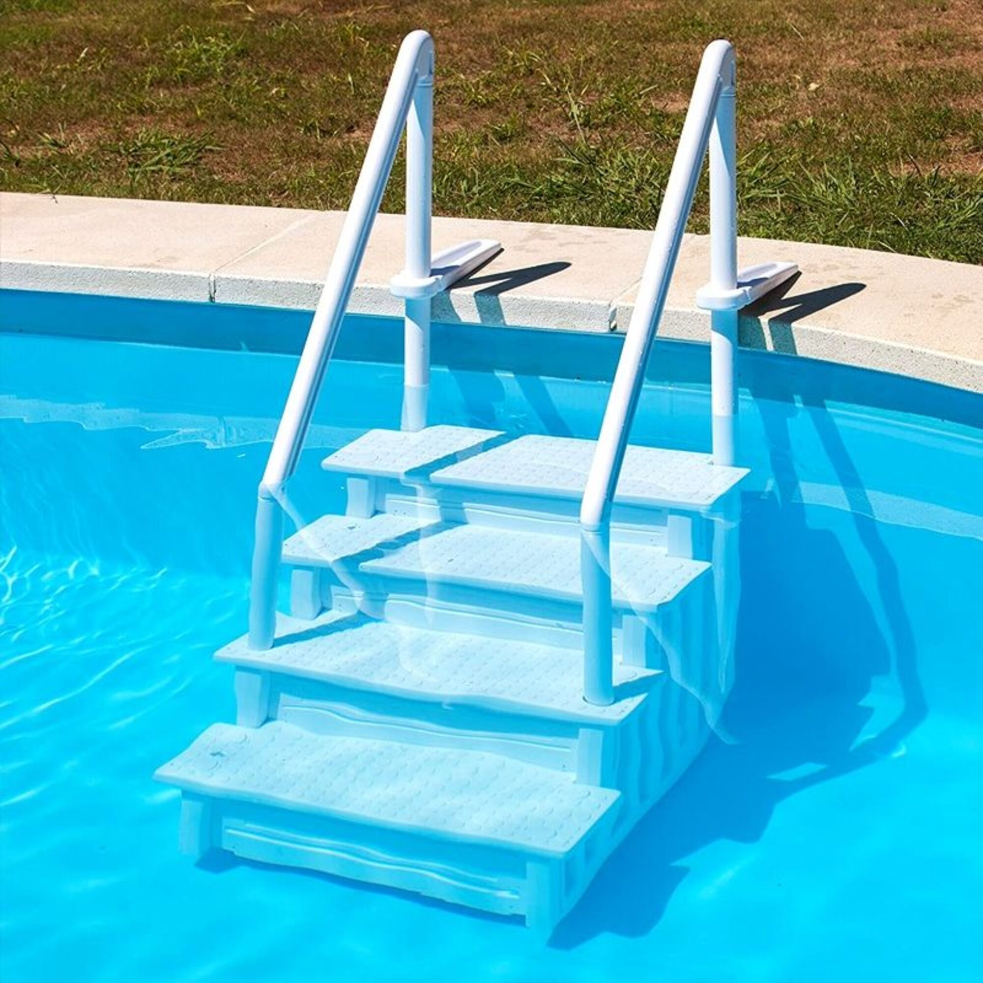Una scala bianca per piscina con gradini è sommersa in acqua blu brillante vicino a un'area erbosa.