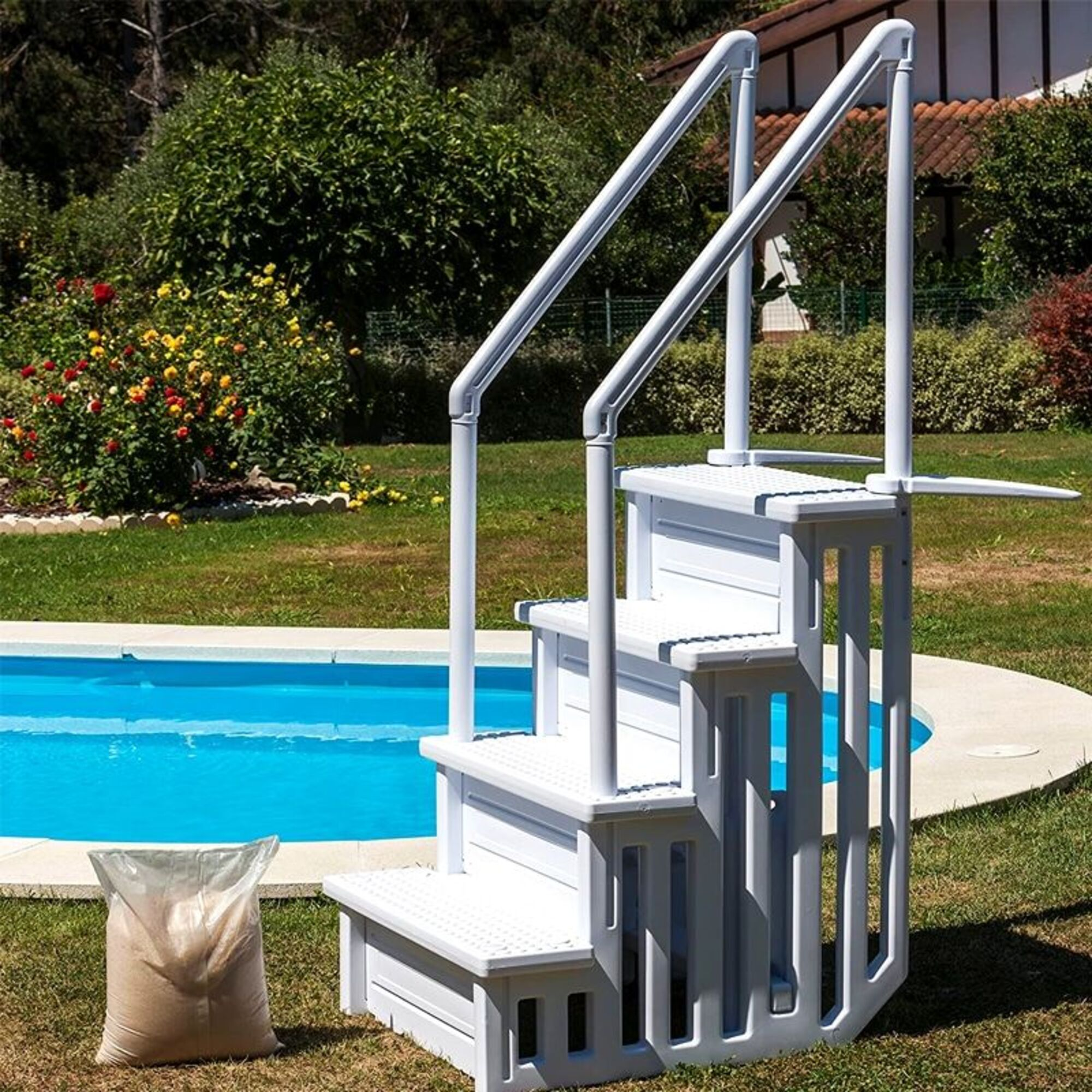 Gradini bianchi per piscina con corrimano accanto a una piscina blu. Un sacco di sabbia sull'erba.