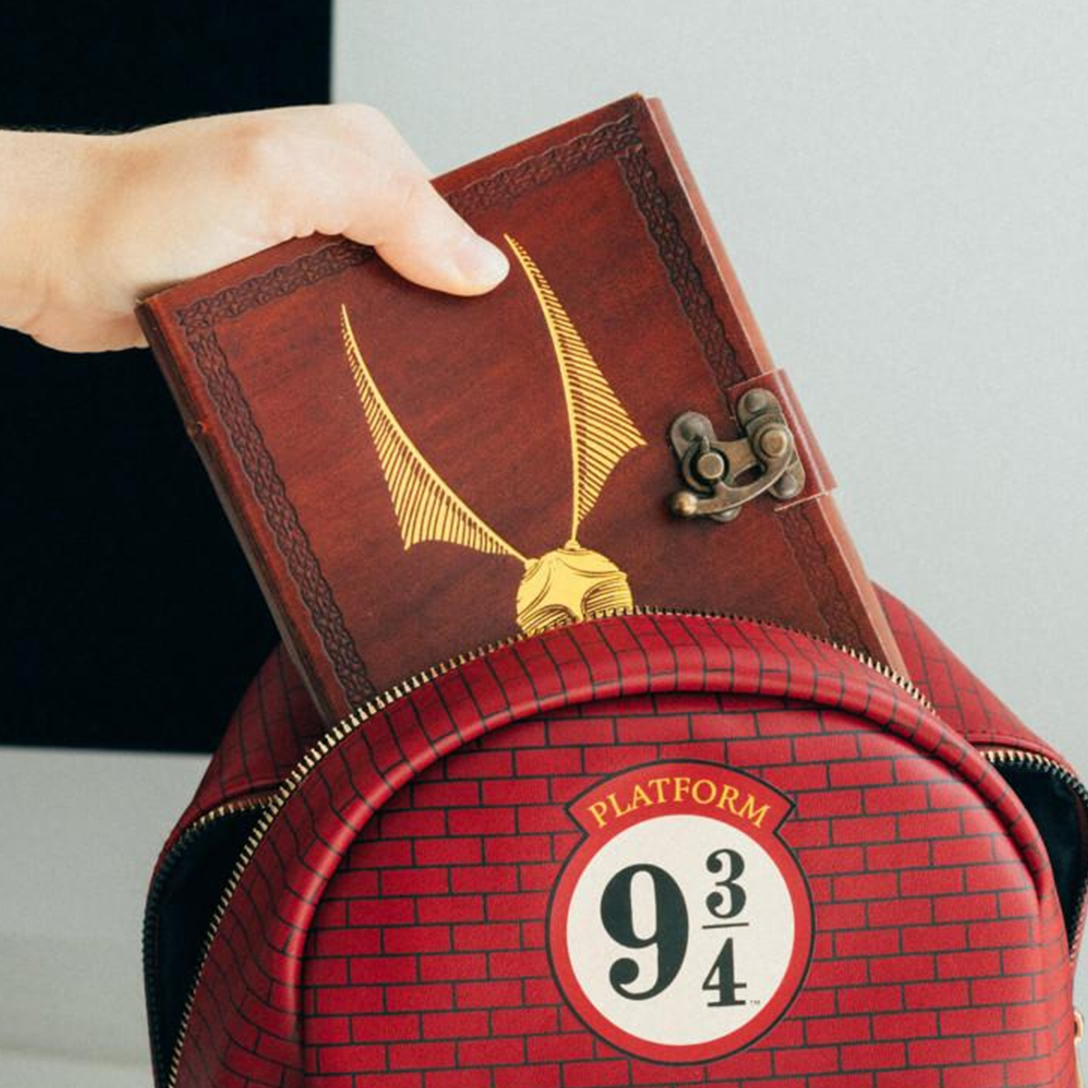 Una mano inserisce un diario del boccino d'oro in uno zaino rosso con il logo del binario 9 3/4.