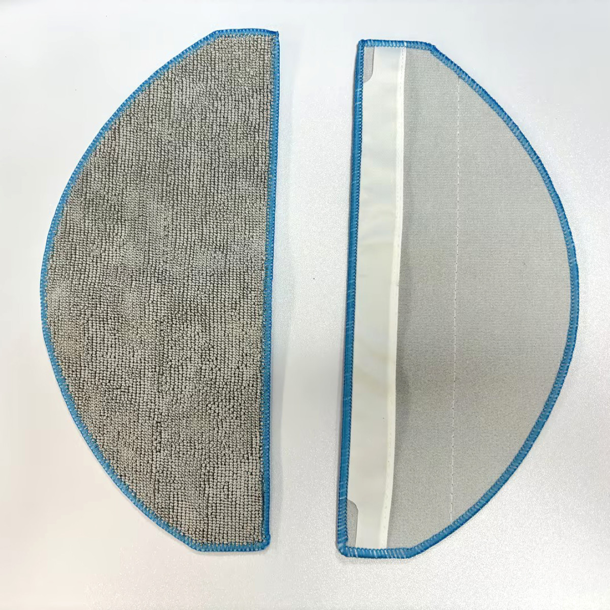 Dwie półokrągłe nakładki czyszczące z niebieskim obszyciem. Jedna to szara mikrofibra, druga to biała siateczka.