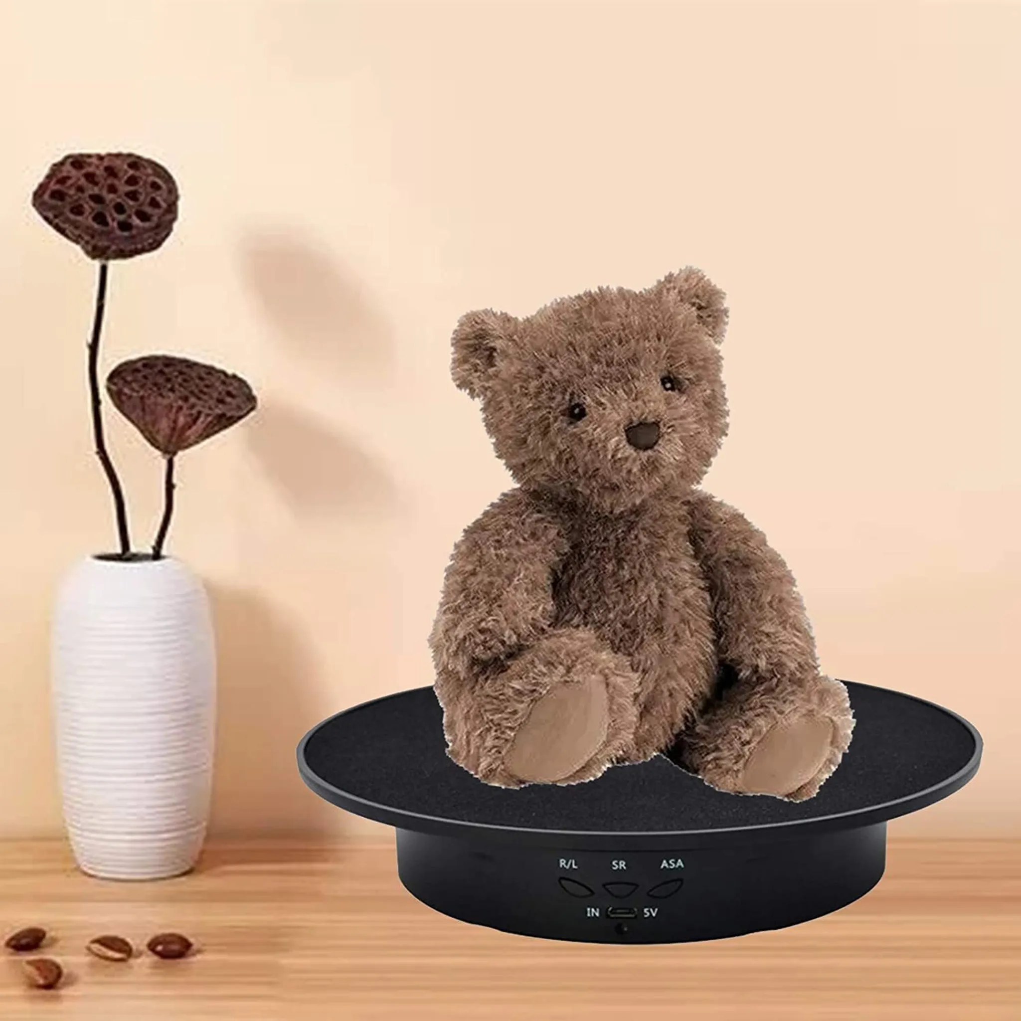 Ein Teddybär sitzt auf einem schwarzen Drehteller neben einer Vase mit getrockneten Lotusstängeln.