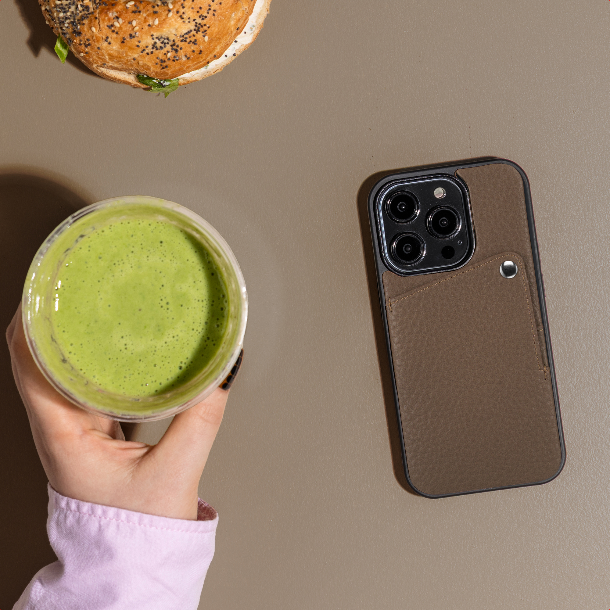 Une main tient un smoothie vert à côté d'un bagel et d'un téléphone avec étui marron.