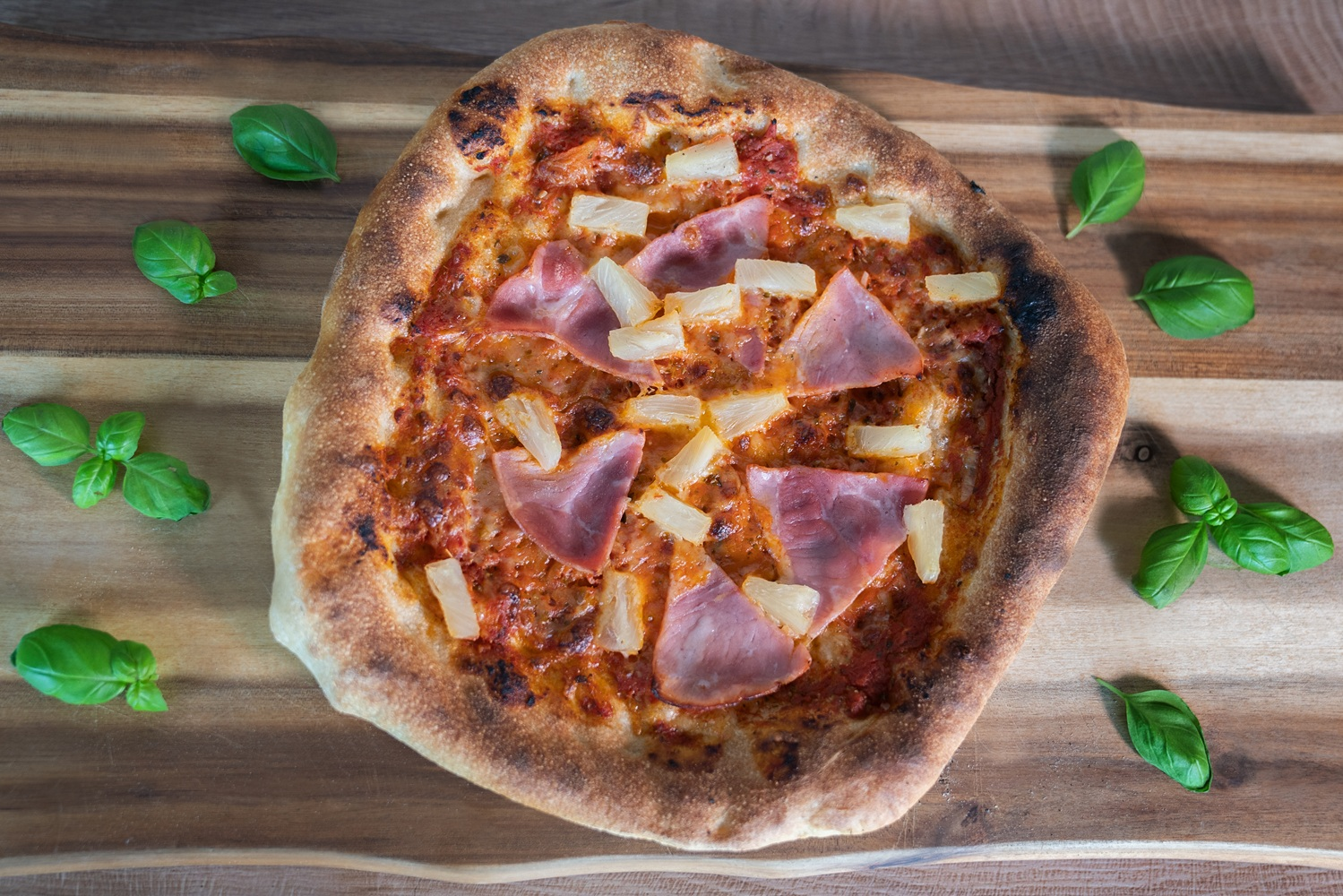 Eine Pizza mit Schinken und Ananas liegt auf einem Holzbrett mit Basilikumblättern.