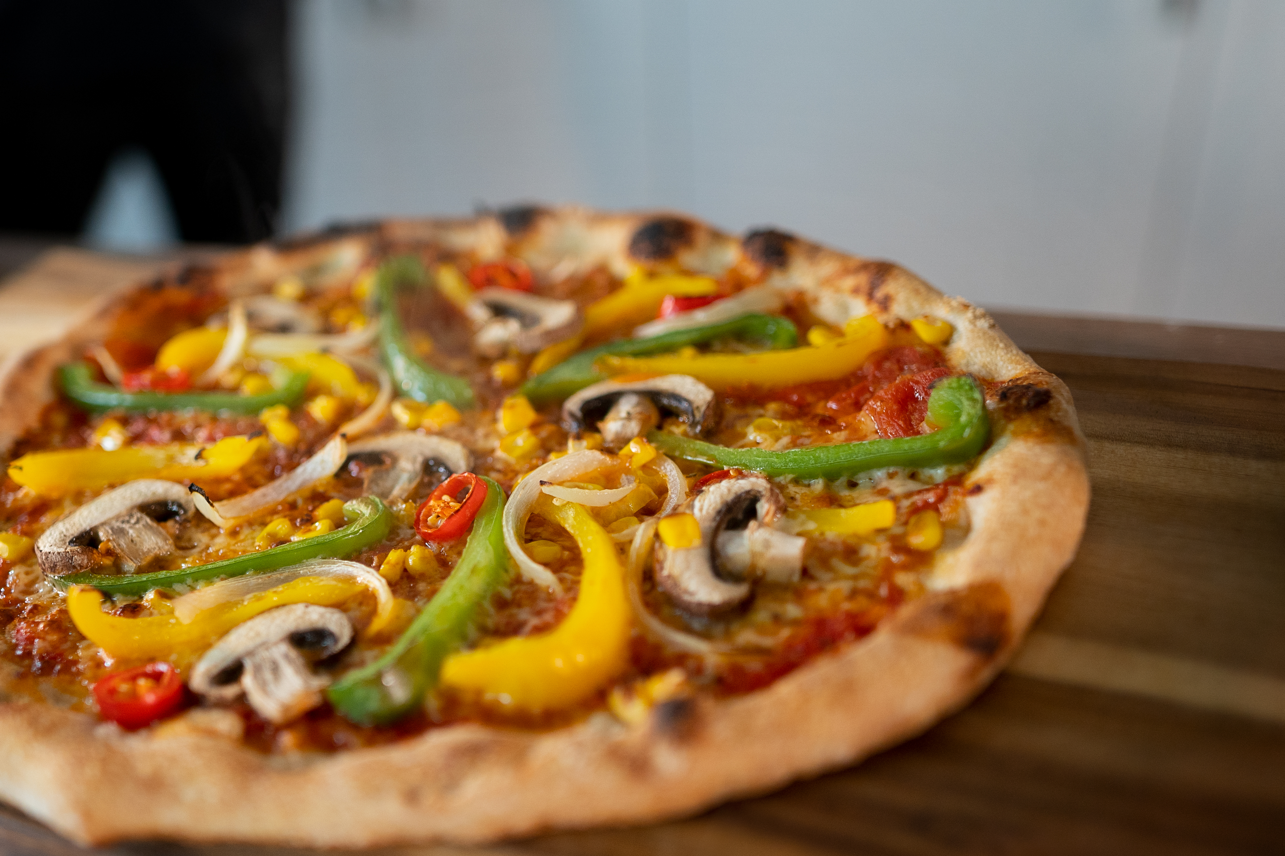 Eine leckere Pizza mit Pilzen, Paprika, Zwiebeln, Mais und Chili auf einem Holzbrett.