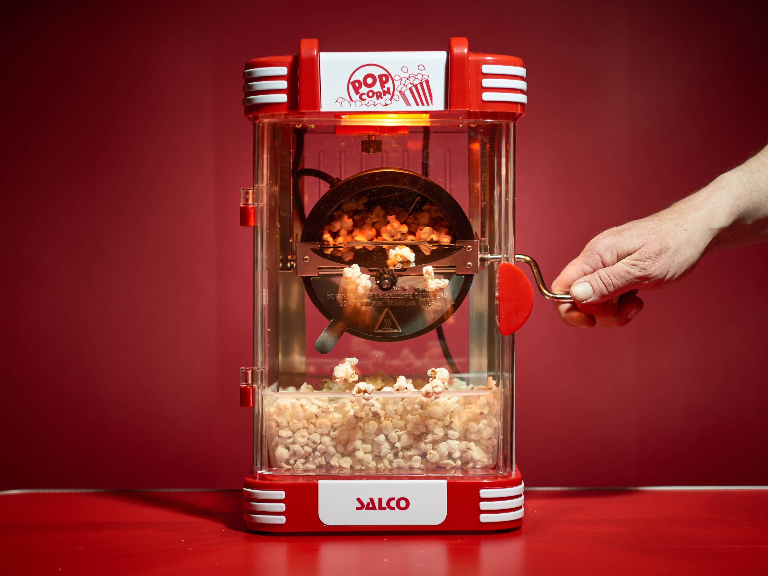 Eine rote Popcornmaschine mit frischem Popcorn darin und einer Hand, die die Kurbel dreht.