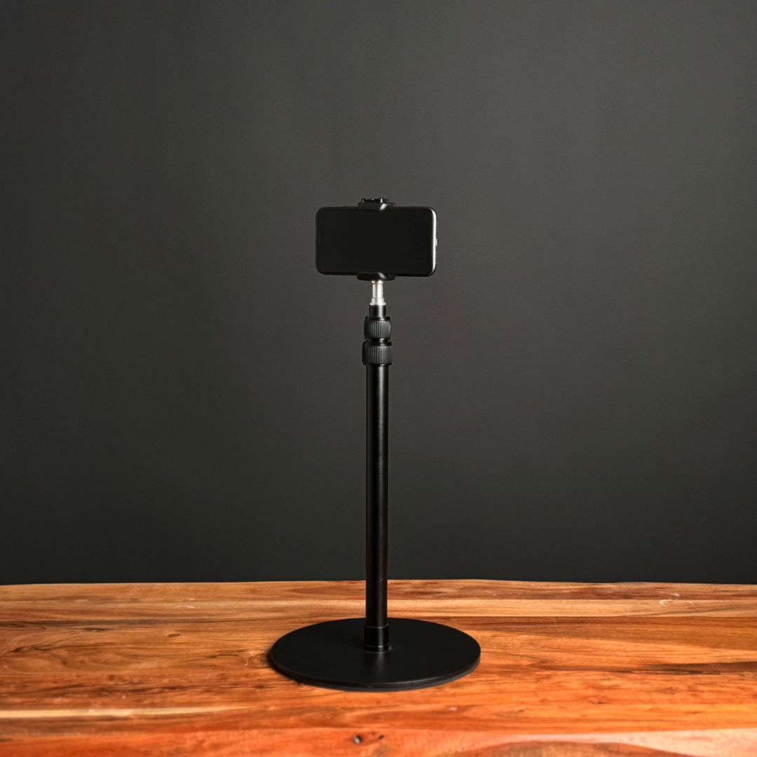 A black smartphone is mounted on a black adjustable stand on a wooden table.