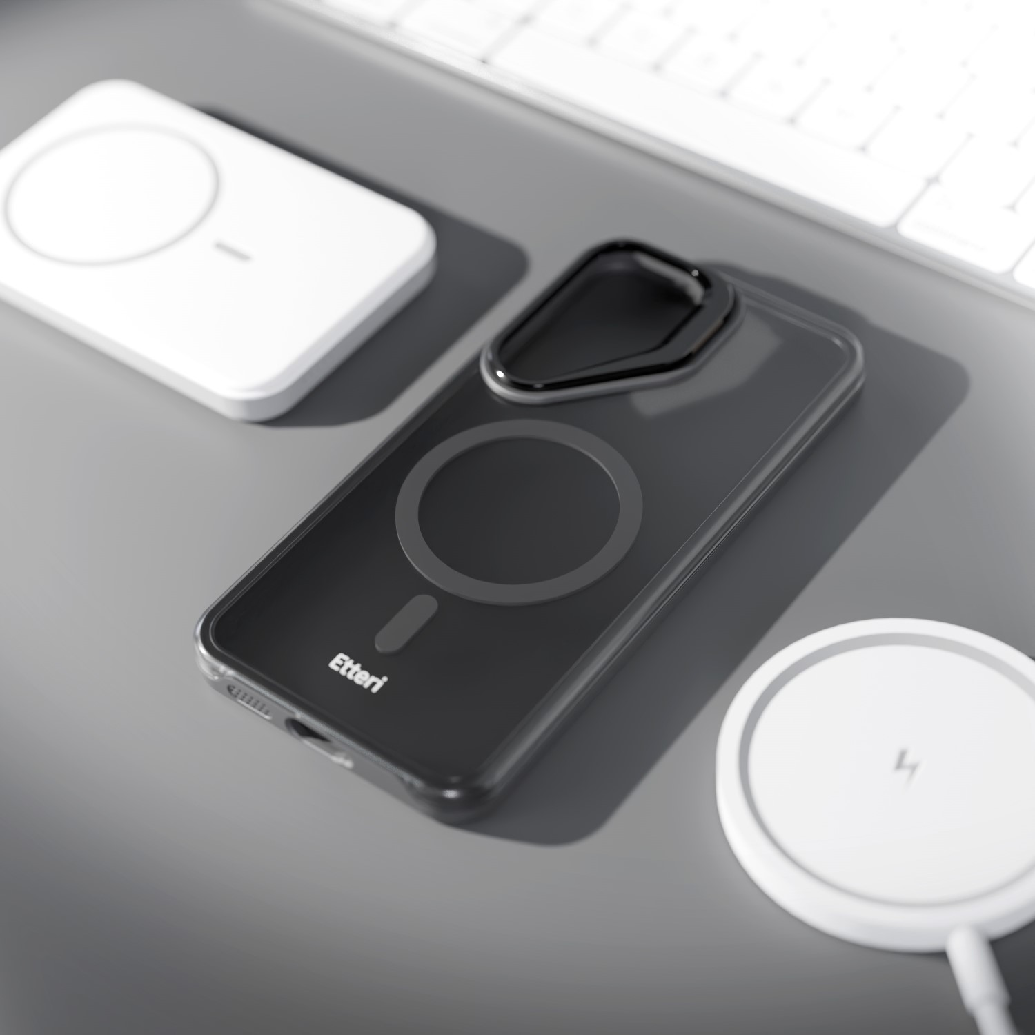Una cover nera per telefono con anello magnetico si trova su una scrivania accanto a due caricabatterie wireless bianchi e una tastiera.
