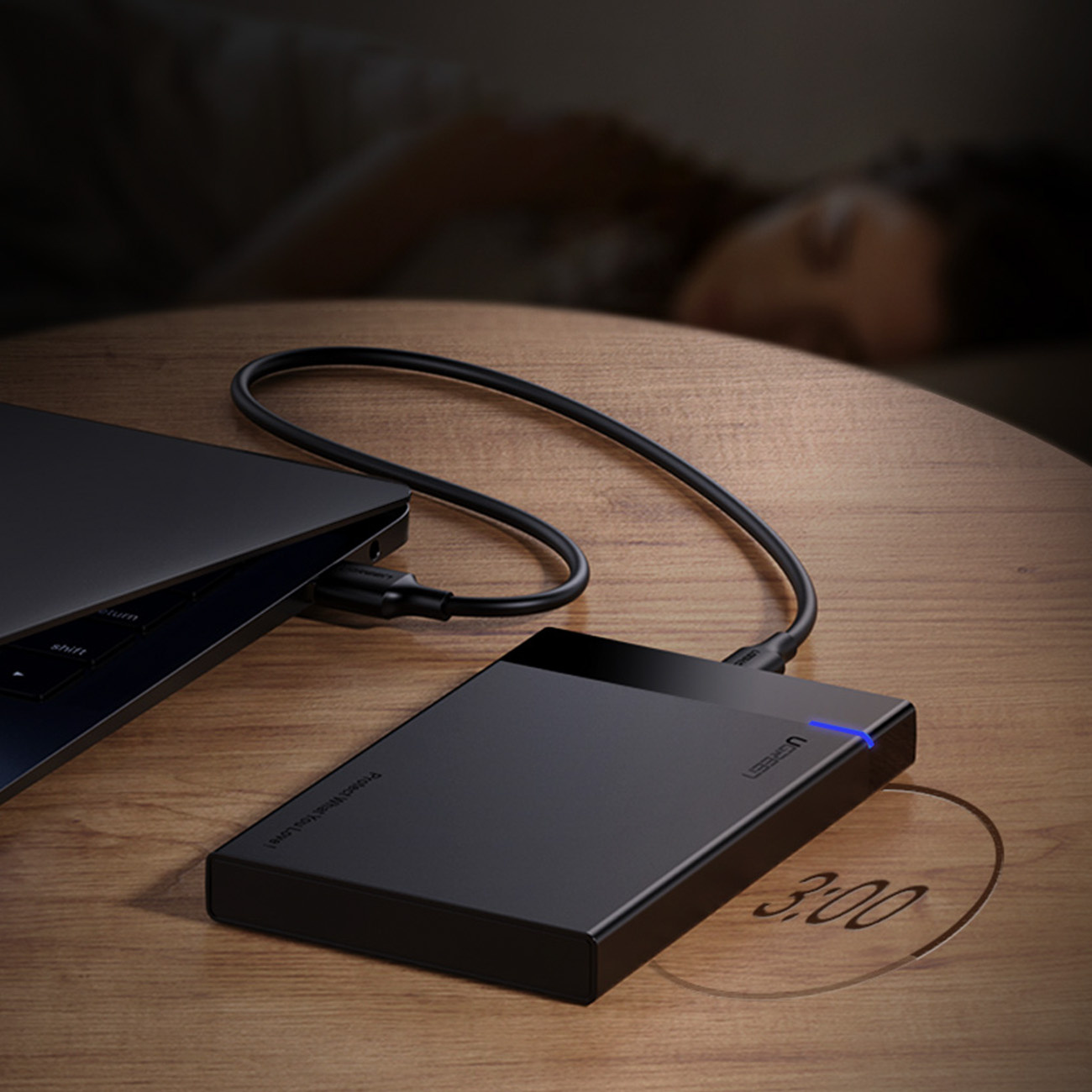 Czarny zewnętrzny dysk twardy podłączony do laptopa na drewnianym stole. W tle śpi osoba.