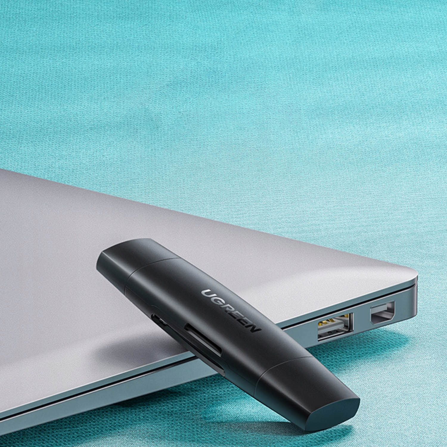 Czarny adapter UGREEN leży na srebrnym laptopie na turkusowym tle.