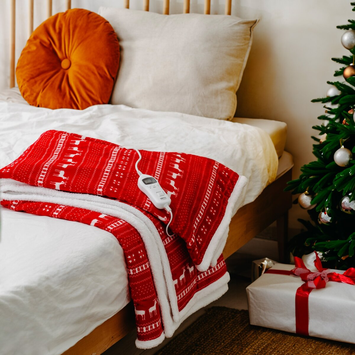 Eine rote Decke mit Rentier-Muster und weißem Rand liegt auf einem Bett neben einem Weihnachtsbaum und Geschenk.