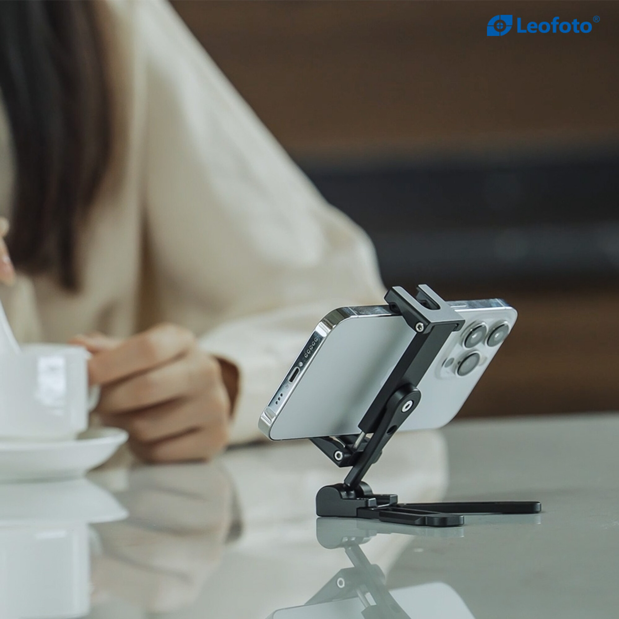 Ein Smartphone auf einem schwarzen Ständer auf einem Tisch. Eine Person hält eine Tasse.