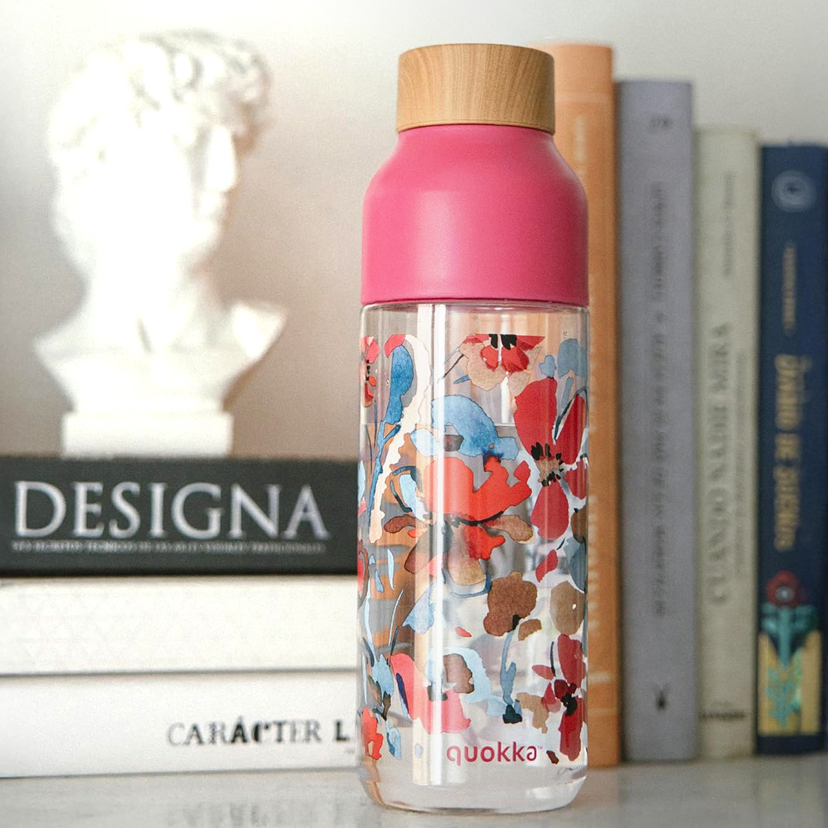 Bottiglia trasparente con fantasia floreale, tappo rosa e coperchio in legno. Libri e busto sullo sfondo.