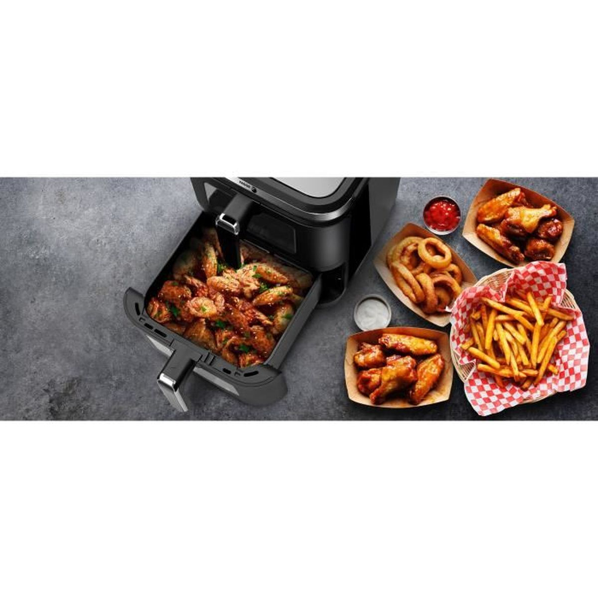 Una friggitrice ad aria con alette di pollo, patatine fritte, anelli di cipolla e ketchup su superficie scura.