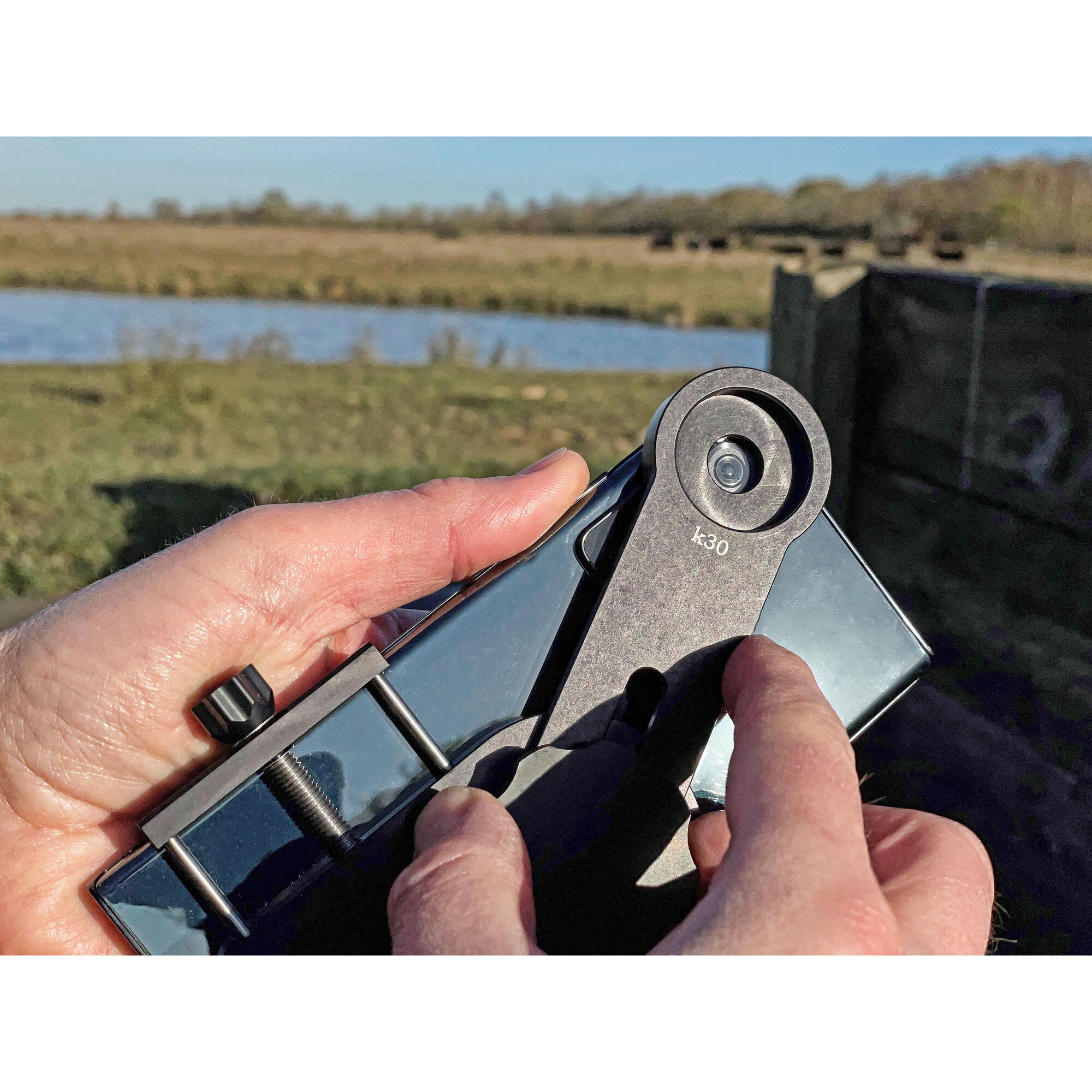 Hände stellen ein Smartphone mit Objektivanbau vor einer Naturlandschaft ein.