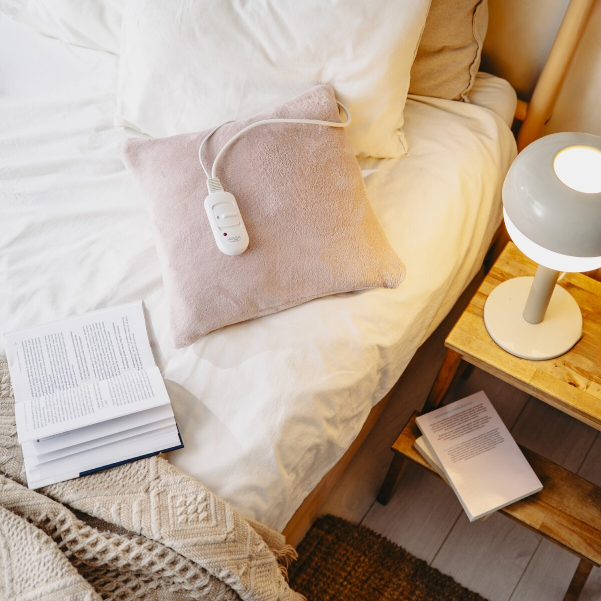 Un letto con lenzuola bianche, un termoforo rosa e un libro aperto. Un comodino contiene una lampada e un altro libro.