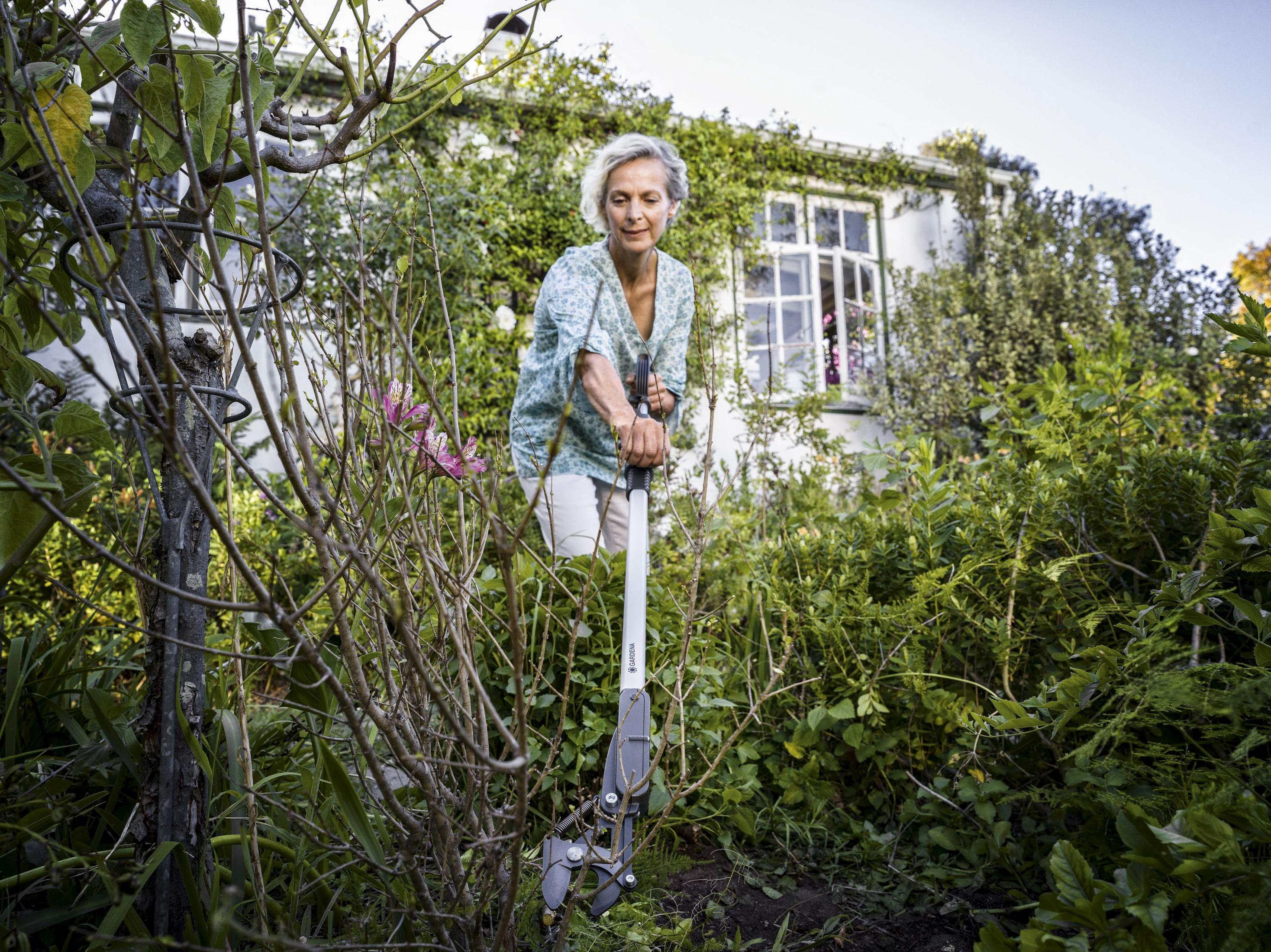 Una donna con i capelli biondi corti pota una pianta con delle cesoie in un giardino lussureggiante.