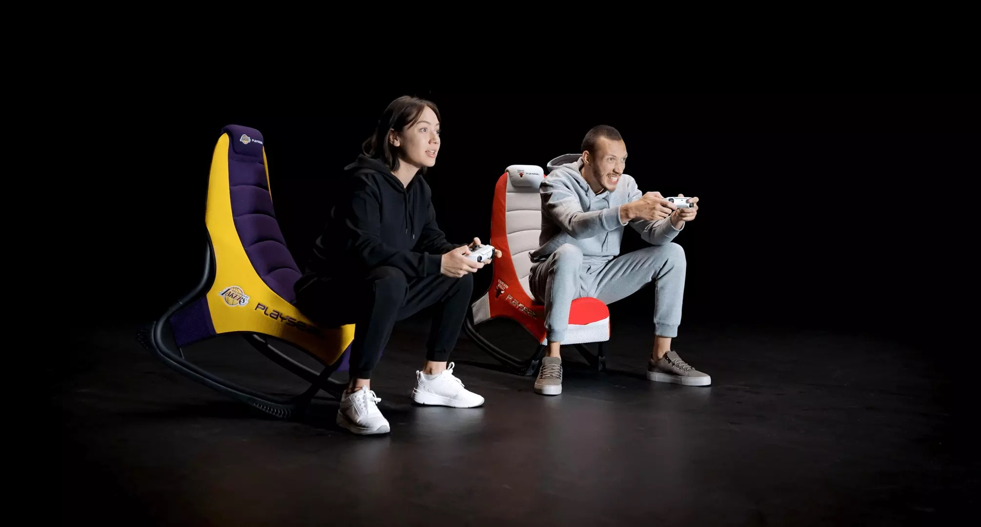 Due persone giocano ai videogiochi su sedie, una nei colori dei Lakers, una in rosso e bianco.