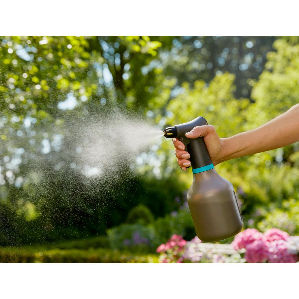 Mano che tiene spruzzino, nebulizzando giardino. Alberi e fiori verdi sullo sfondo.
