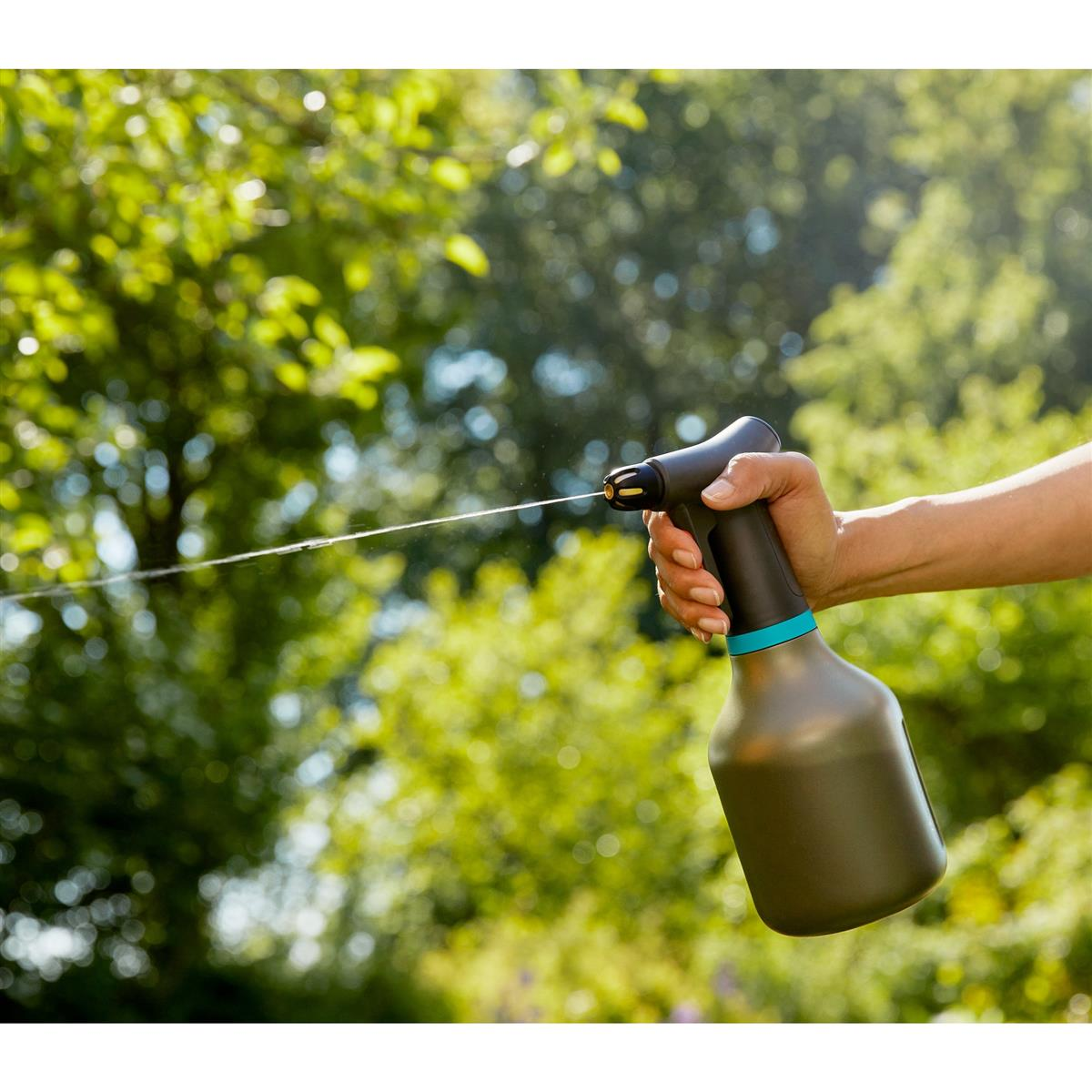 Mano tiene una bottiglia spray grigio scuro con anello blu, spruzza acqua all'aperto con alberi verdi.