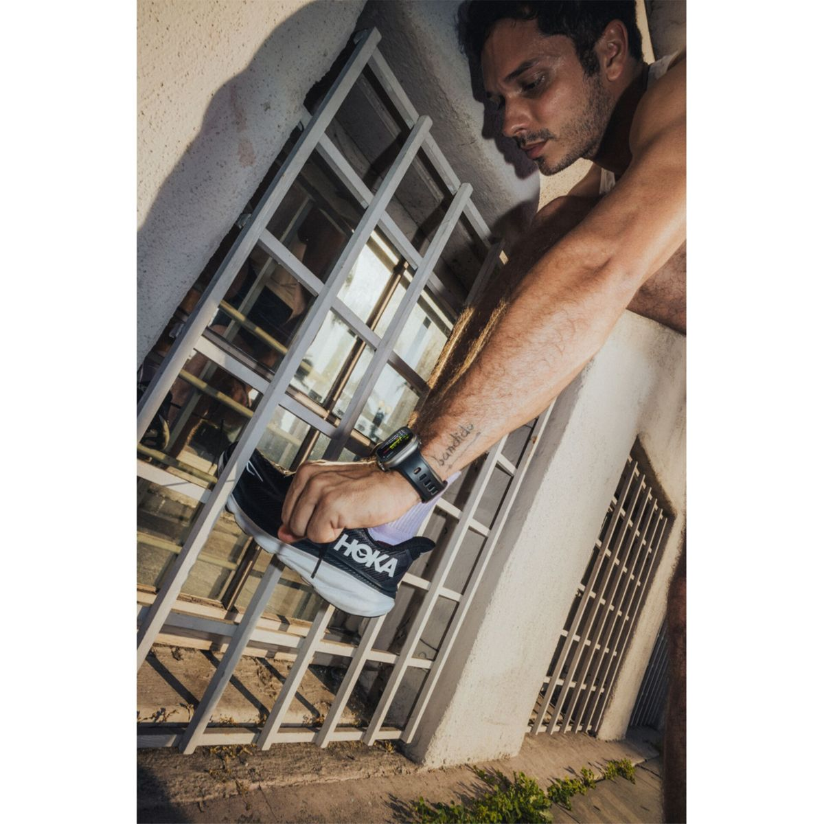 Hombre se pone zapatillas Hoka cerca de rejas de ventana. Lleva reloj y tatuaje.