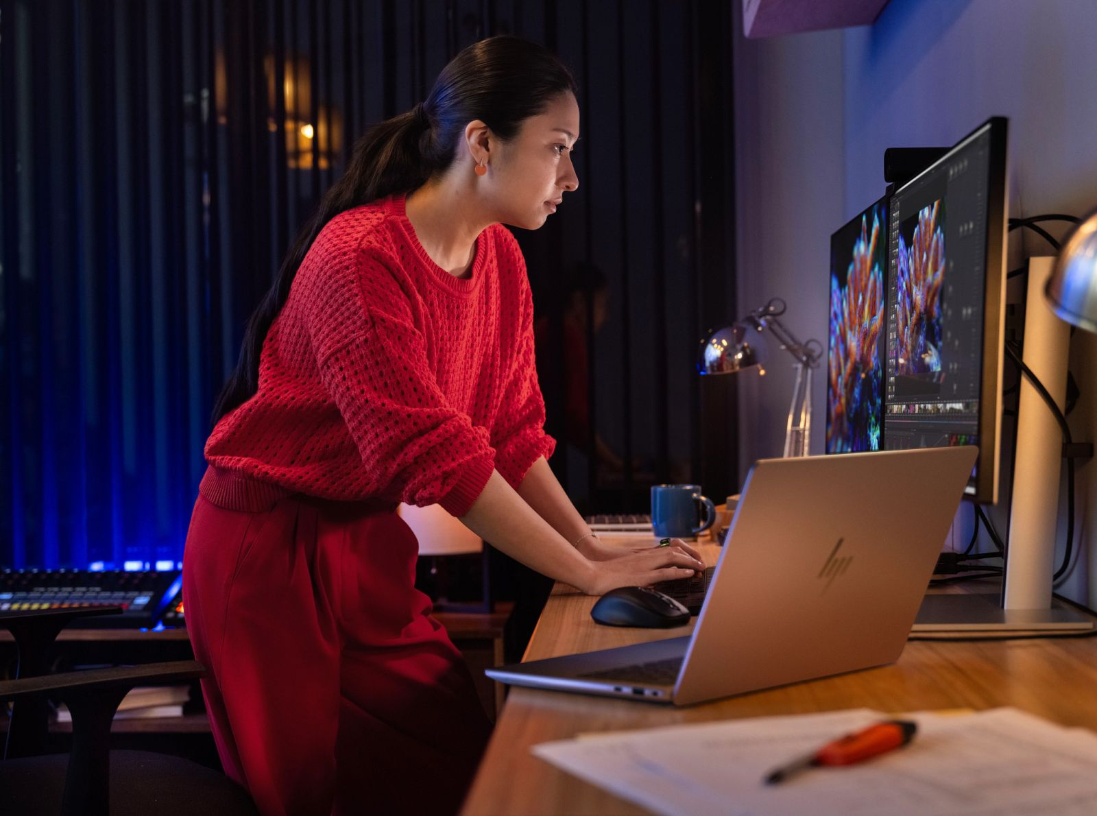 Kobieta w czerwonym swetrze pracuje przy laptopie i dwóch monitorach późno w nocy.