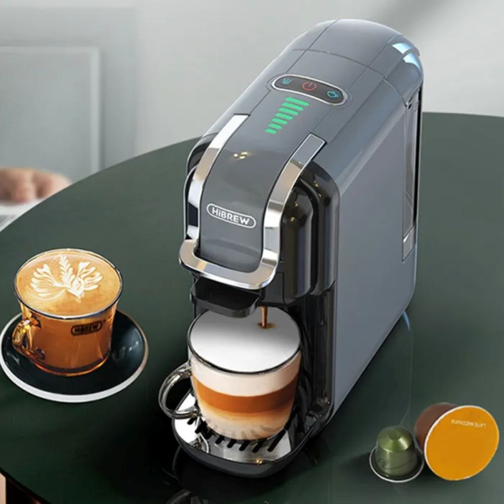 Una cafetera gris prepara un latte en una taza de cristal, con otro latte y cápsulas de café cerca.