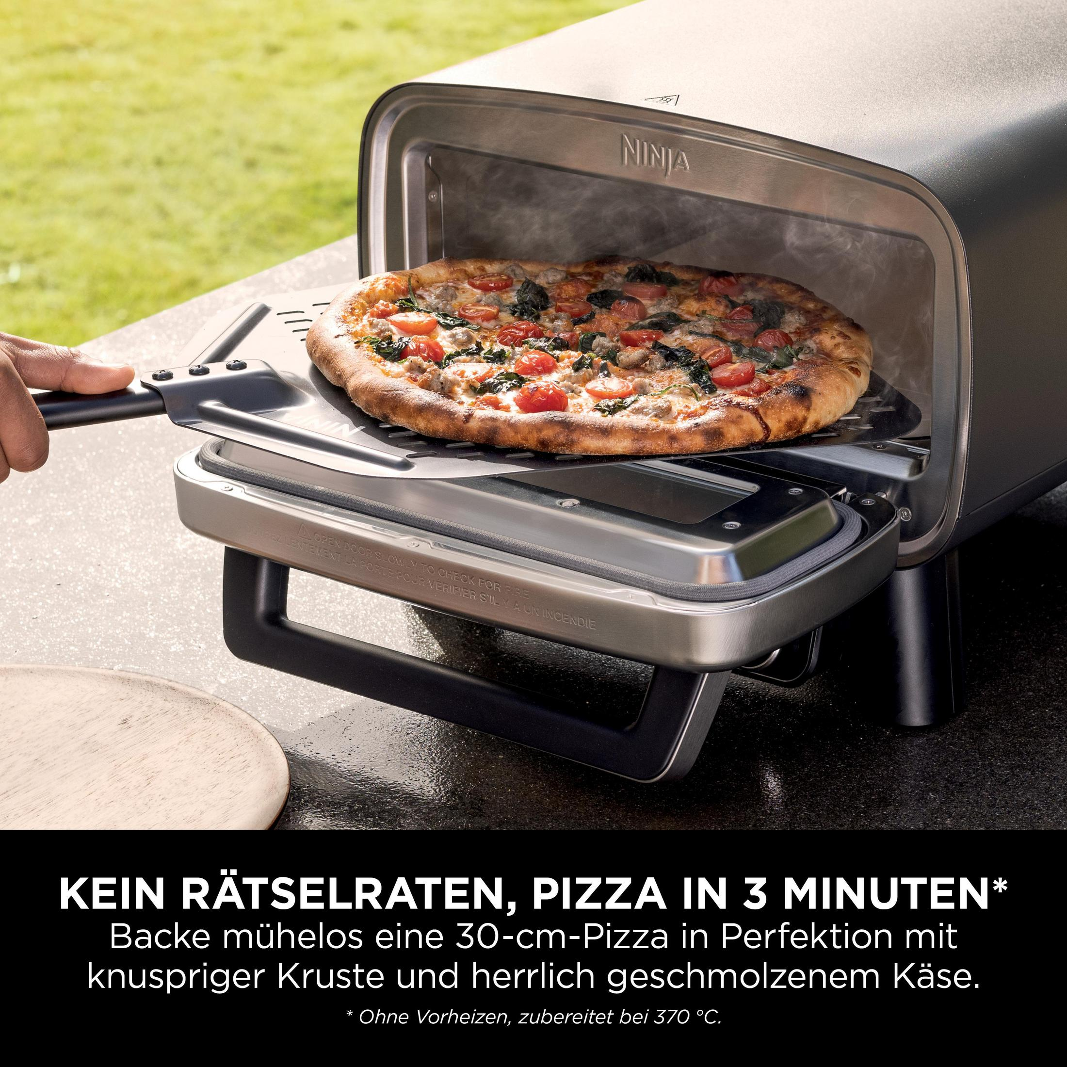 A hand places a pizza on a pizza peel into a Ninja oven outdoors. Blurred grass background.