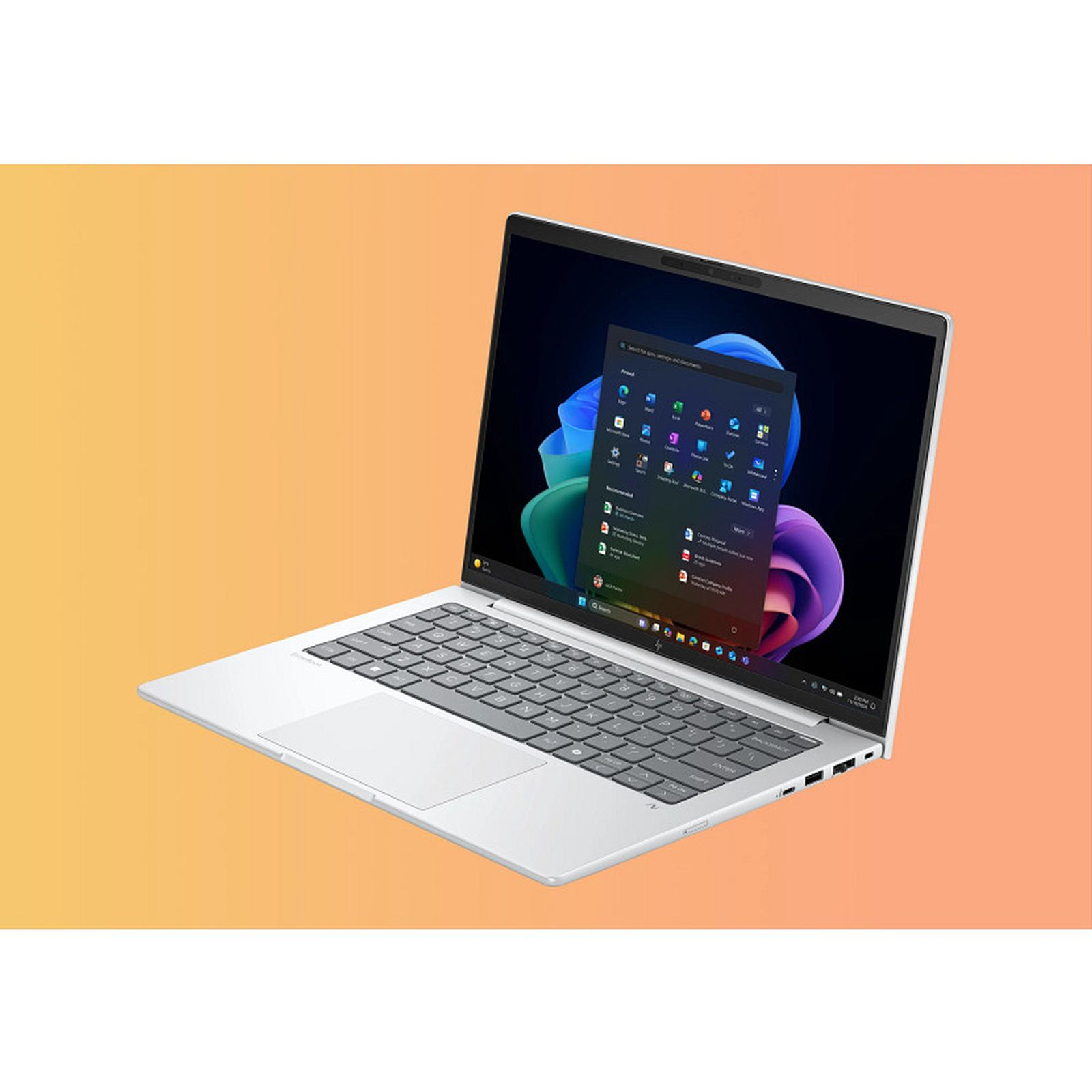 Srebrny laptop z otwartym menu startowym Windows 11 na ekranie. Pomarańczowe tło.
