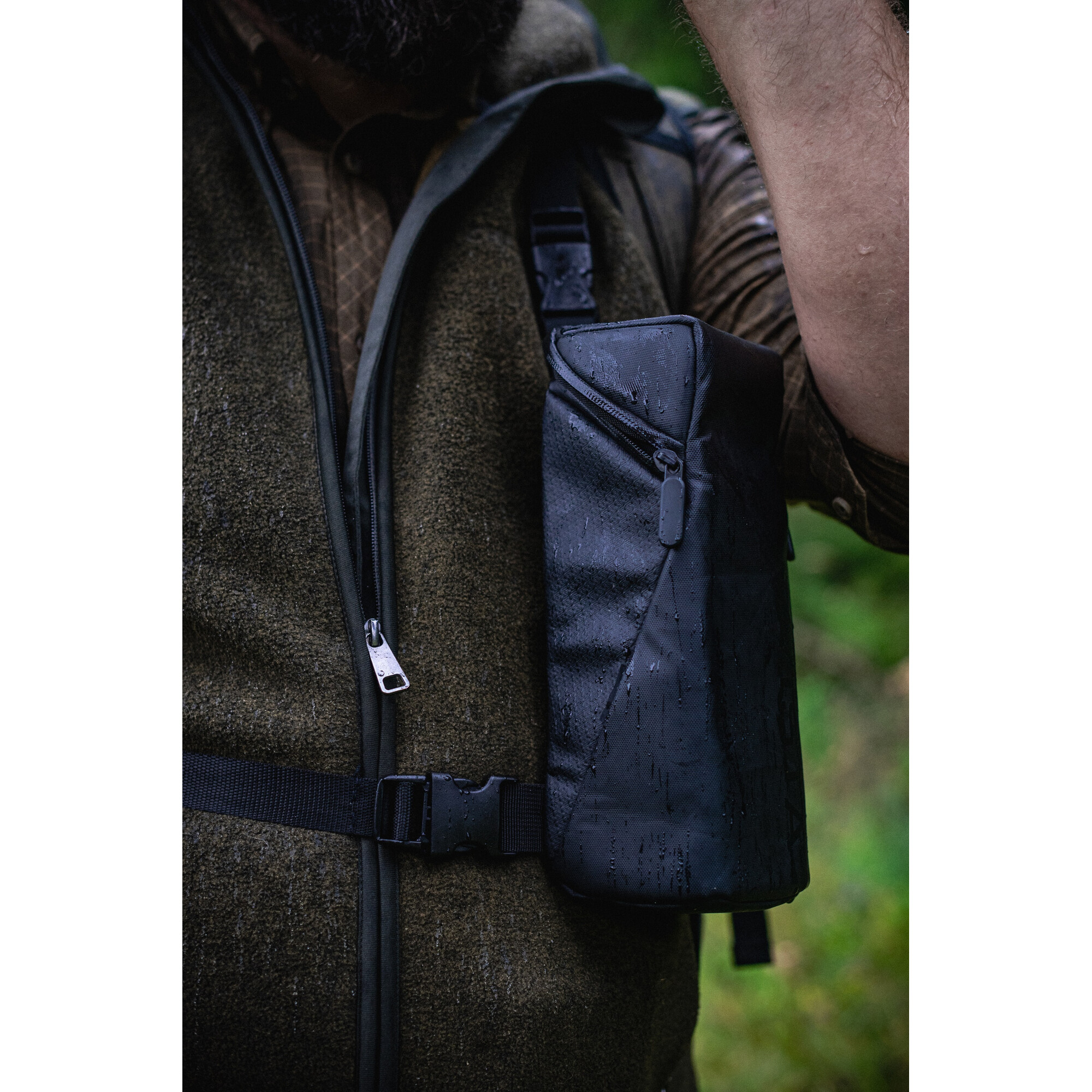 Eine Person mit Weste und Rucksack im Wald. Der Rucksack hat Wassertropfen.