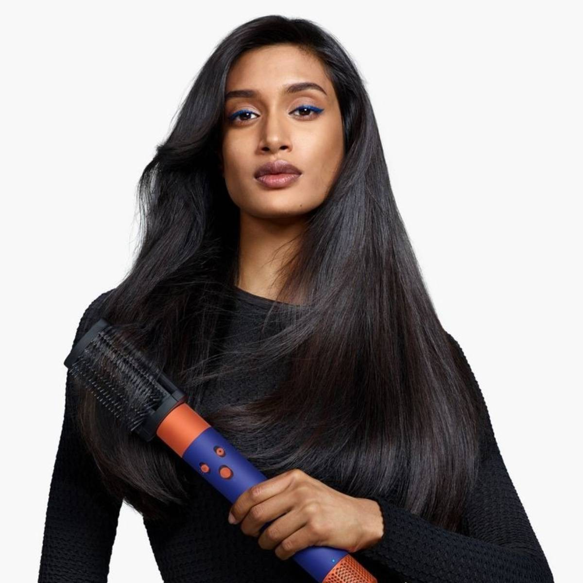 Donna con lunghi capelli scuri e eyeliner blu tiene una spazzola asciugacapelli blu e arancione.